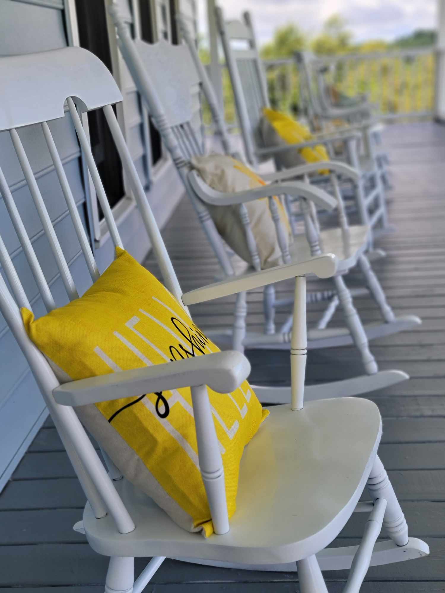 White rocking chairs with yellow pillows on a porch, facing outward with a view of trees and sky.