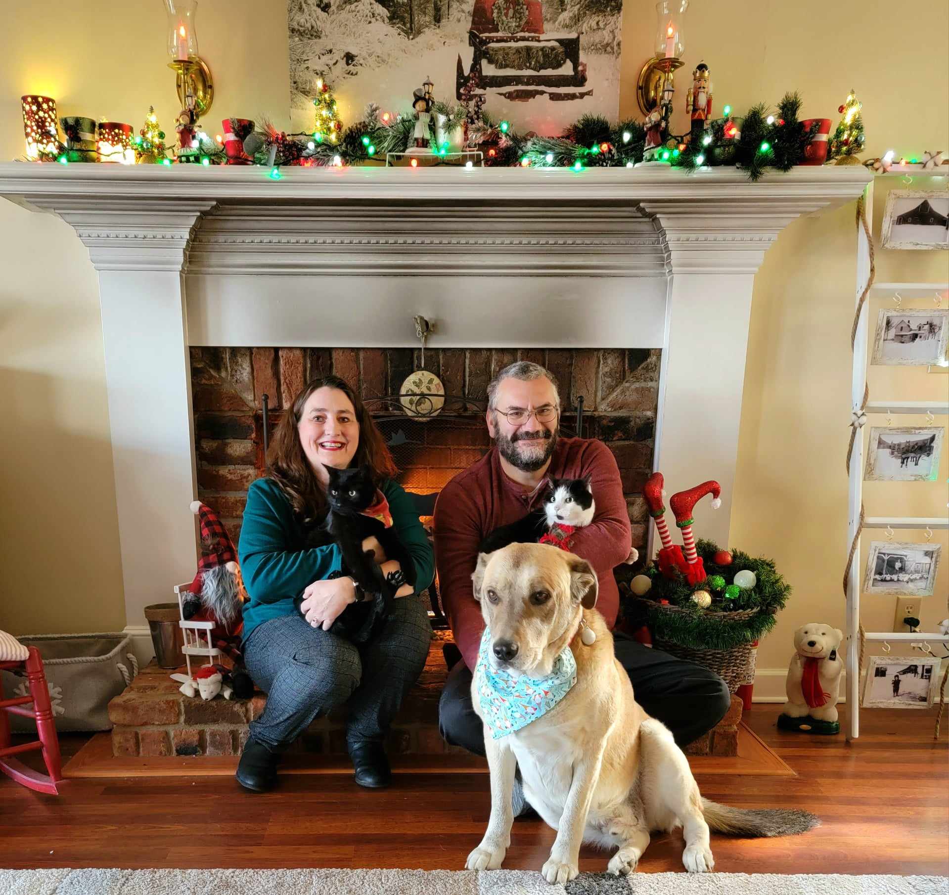 Two people, a dog, and a cat in front of a decorated fireplace with Christmas decorations.