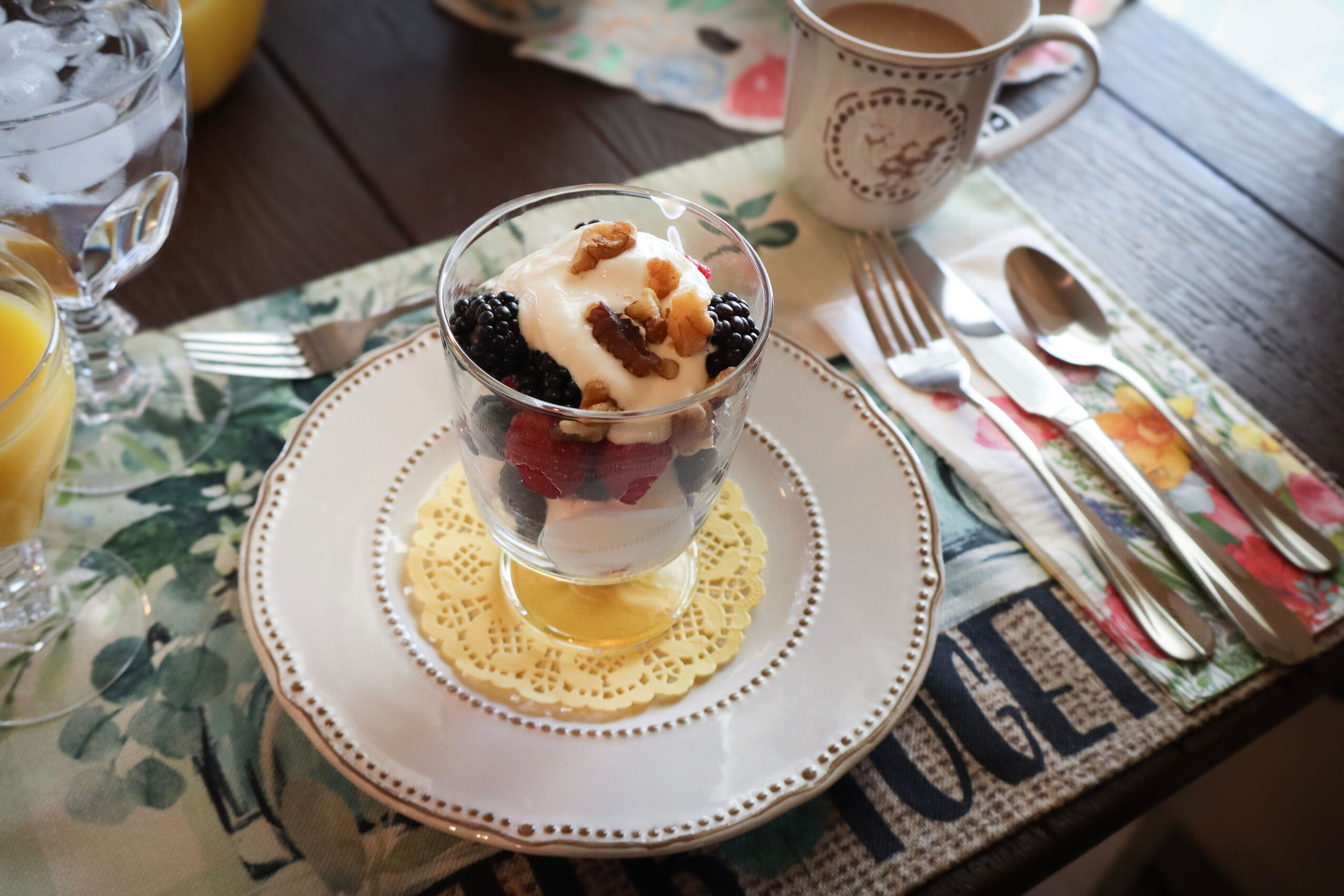Dessert in a glass with berries and cream on a decorative plate, set on a table with utensils and a cup.