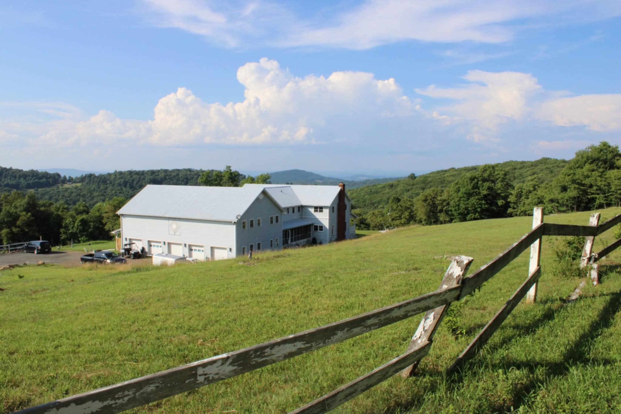 A white barn on a grassy hill with a wooden fence, green trees, and a blue sky with clouds.