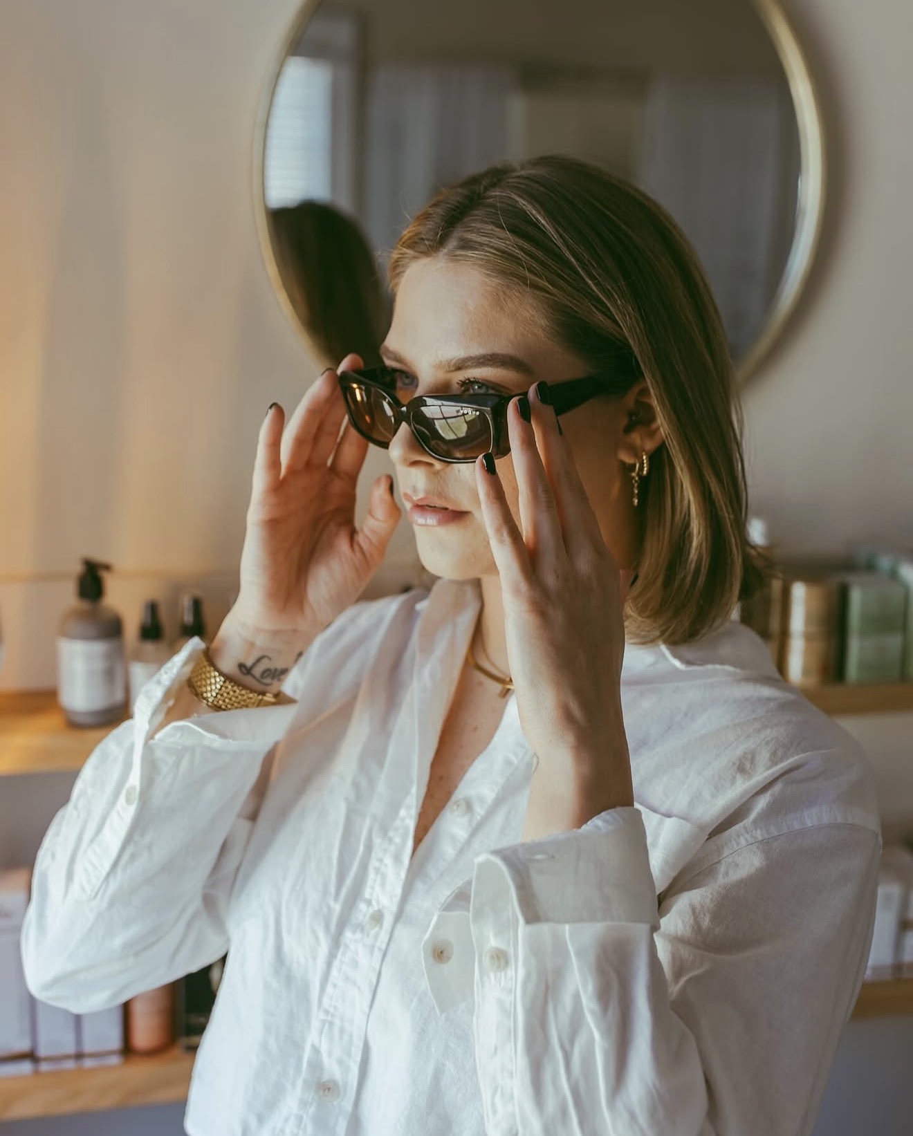 Woman adjusting sunglasses in front of a mirror, wearing a white shirt, with a blurred background of bottles and containers.