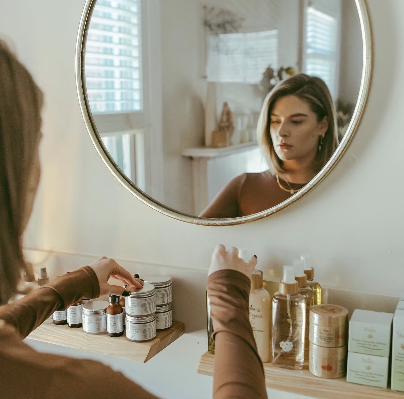 Woman looking at her reflection in a round mirror, with skincare products on a table, in a bright room.