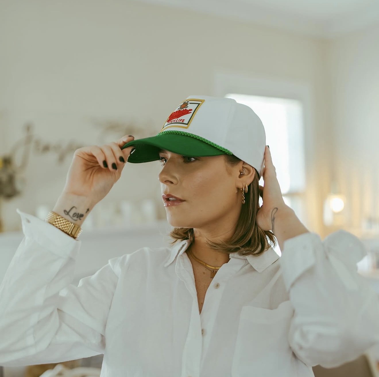 Woman wearing a white shirt and a white cap with a green brim, adjusting the cap in a bright room.