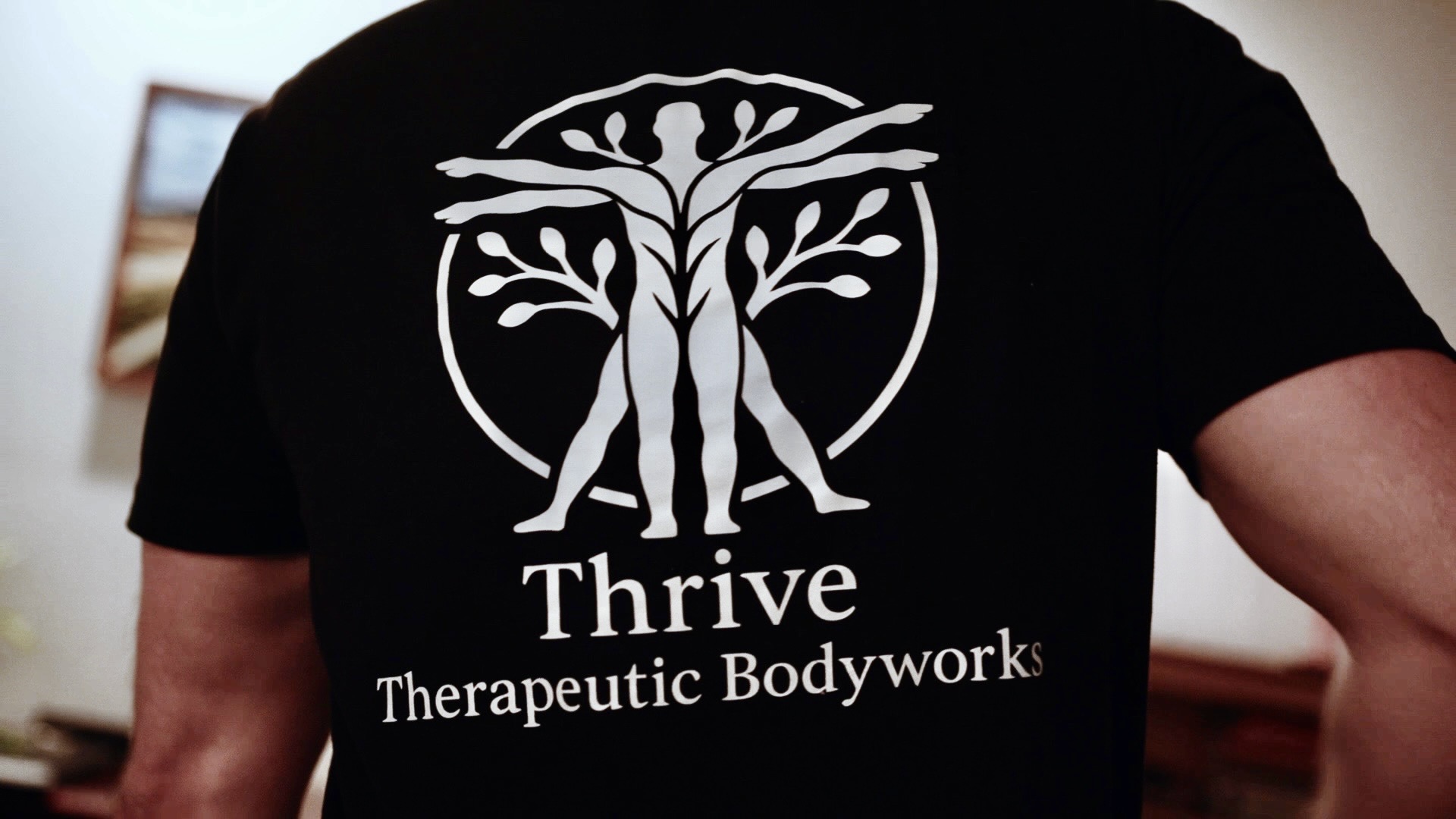 Person wearing a black T-shirt with a white tree logo and text, standing indoors.