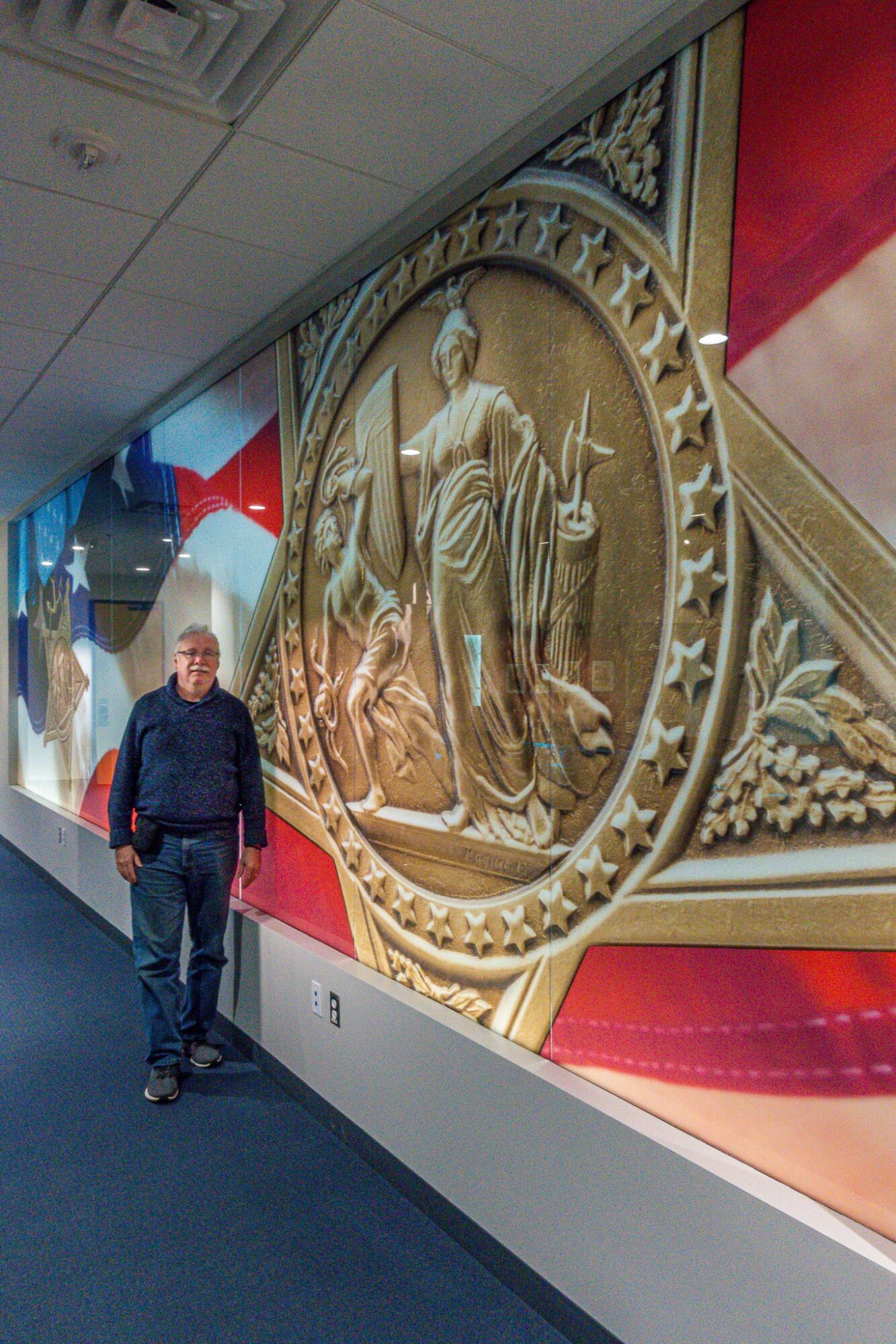 Man walking in hallway with large circular decorative artwork on wall featuring a figure holding a staff, surrounded by ornate details.
