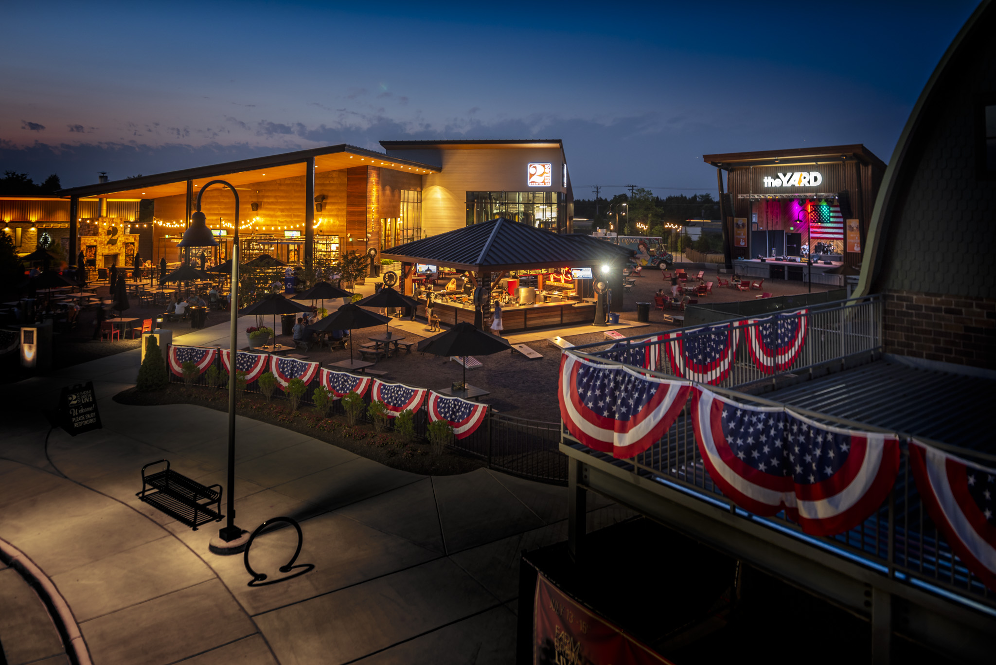 Nighttime scene of a lively outdoor area with decorated bunting, illuminated buildings, and a stage or performance space.