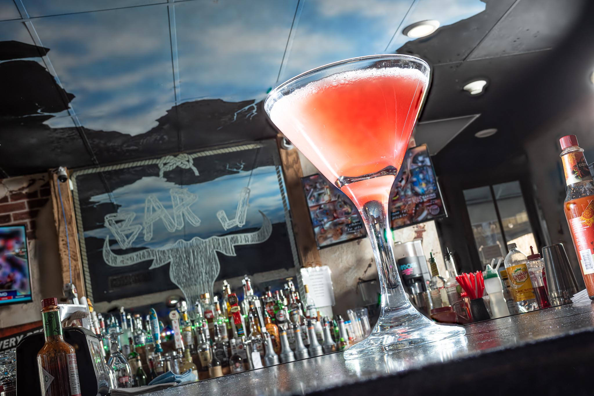 Pink cocktail in a martini glass on bar counter with bottles and bar equipment in background