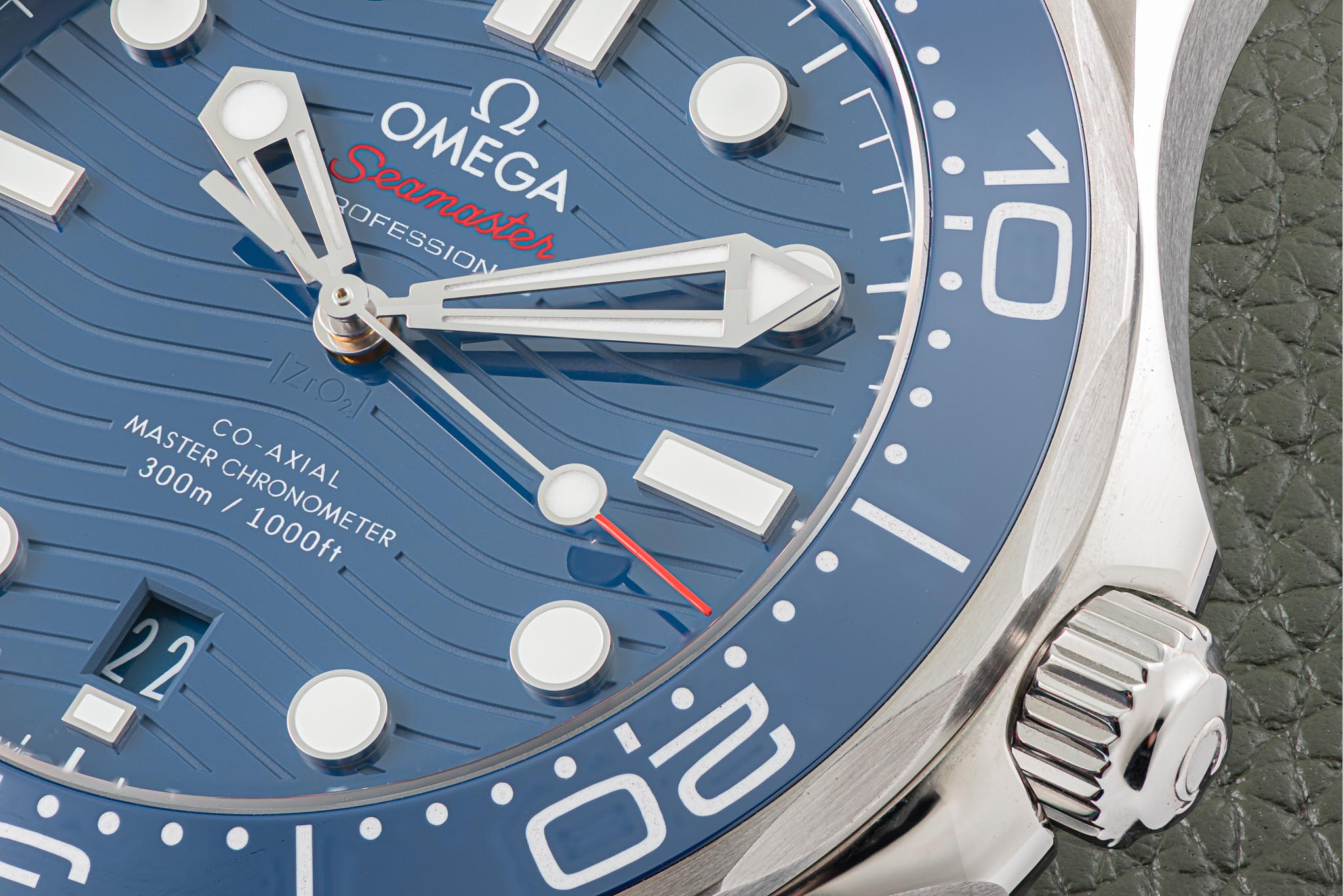 Close-up of a blue Omega Seamaster watch face with white markers and hands, and a textured green strap visible on the right.