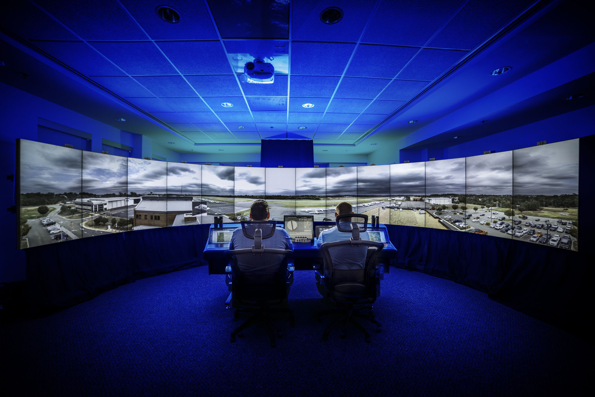 Two people sit at a control desk with multiple large screens displaying a cityscape, in a dark room with blue lighting.