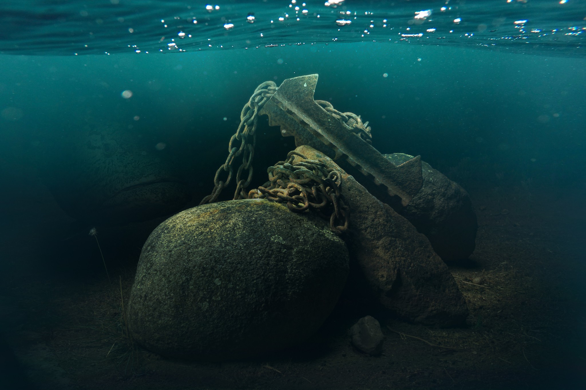 Underwater scene with large rocks and a chain attached to a submerged object, water visible at the surface.