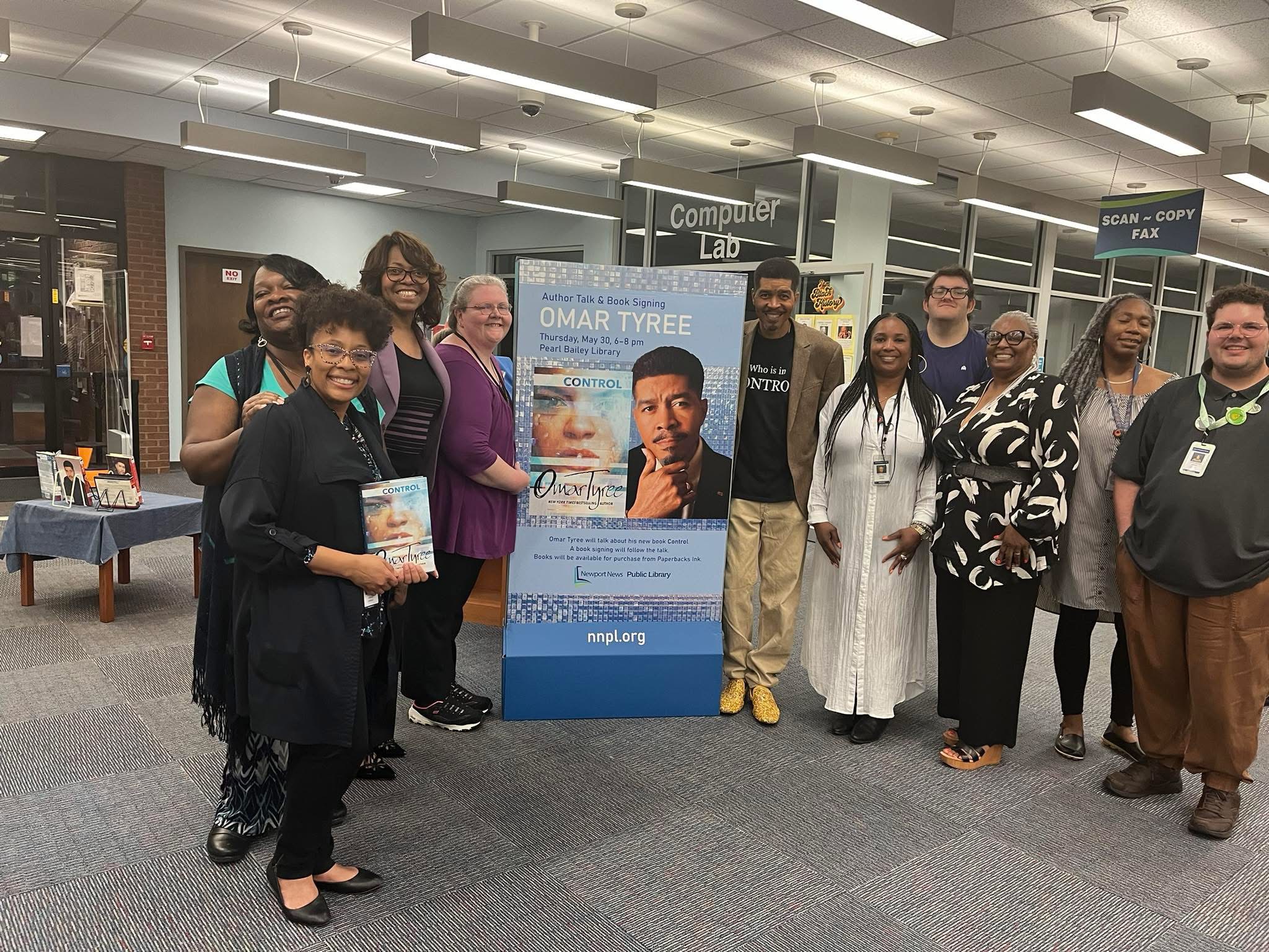 Group of ten people standing indoors around a poster of a man, some holding books, in a room with computer lab signs.
