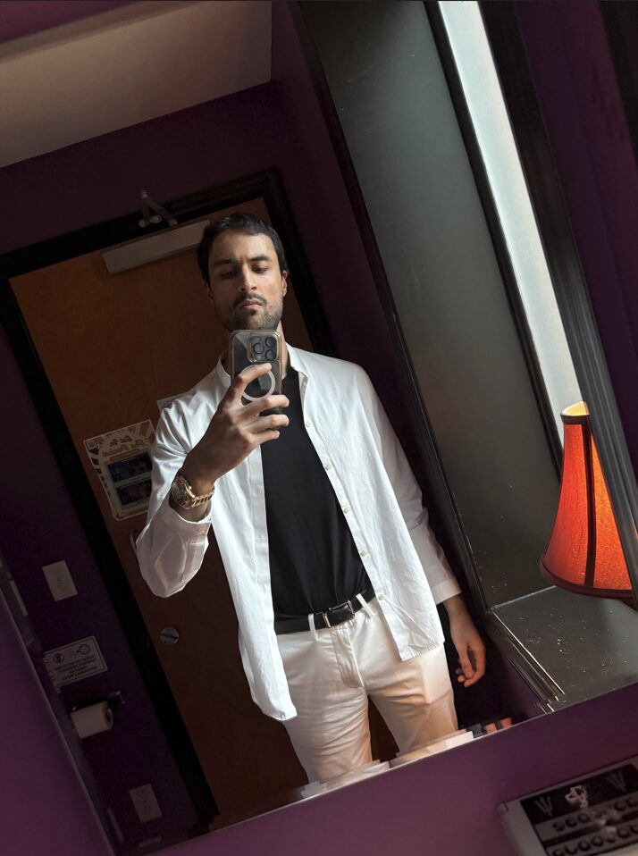 Man taking a mirror selfie in a room with purple walls, wearing a white jacket, black shirt, and beige pants.
