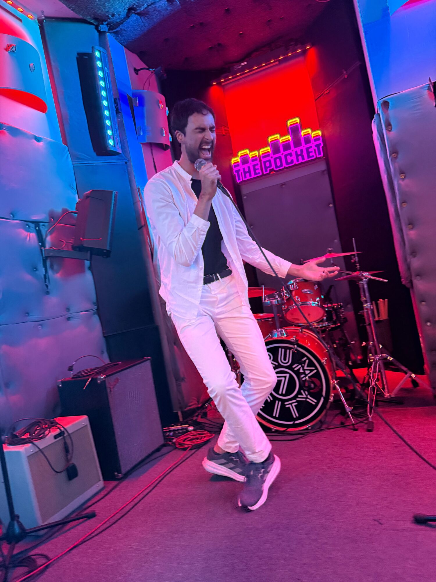 Person in white outfit standing in a room with colorful neon lights, holding a peace sign, with DJ equipment nearby.