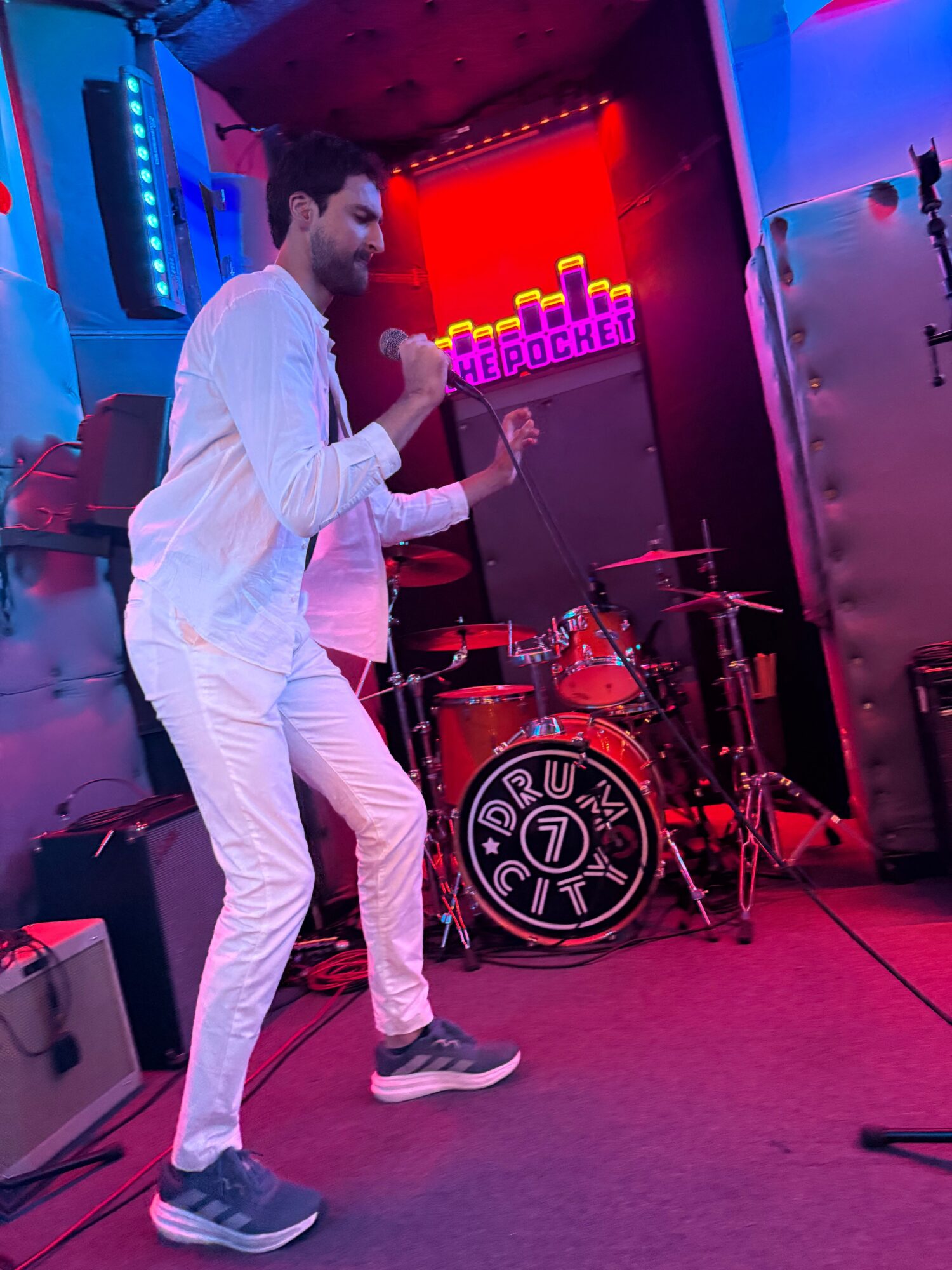 Person singing into a microphone on stage with drums and colorful lighting, wearing white clothing and sneakers.
