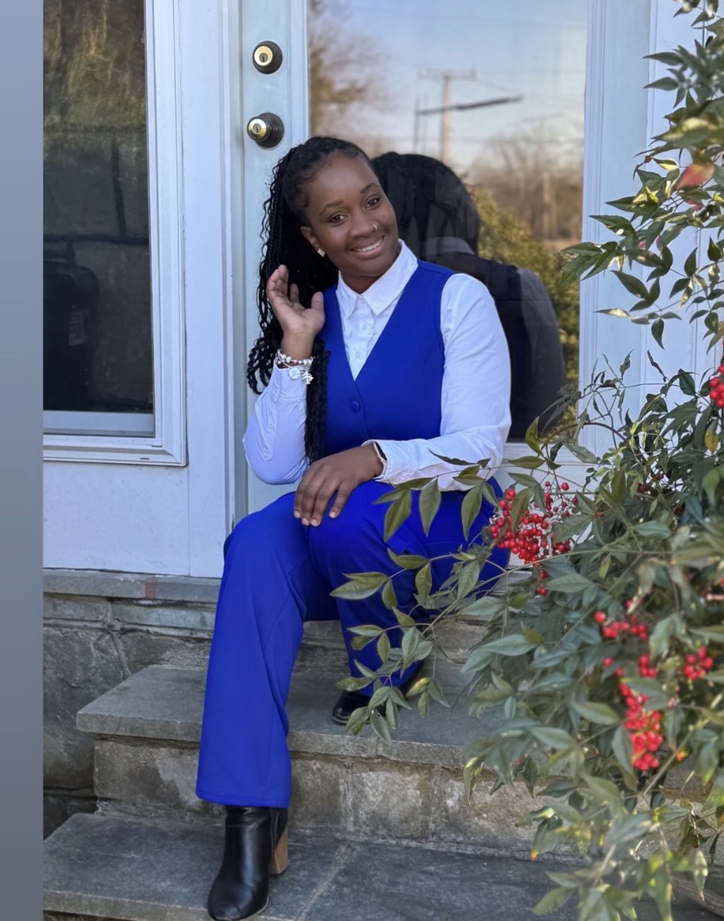 Woman sitting on steps outside, wearing blue pants and vest, white shirt, smiling, with plants and a door behind her.