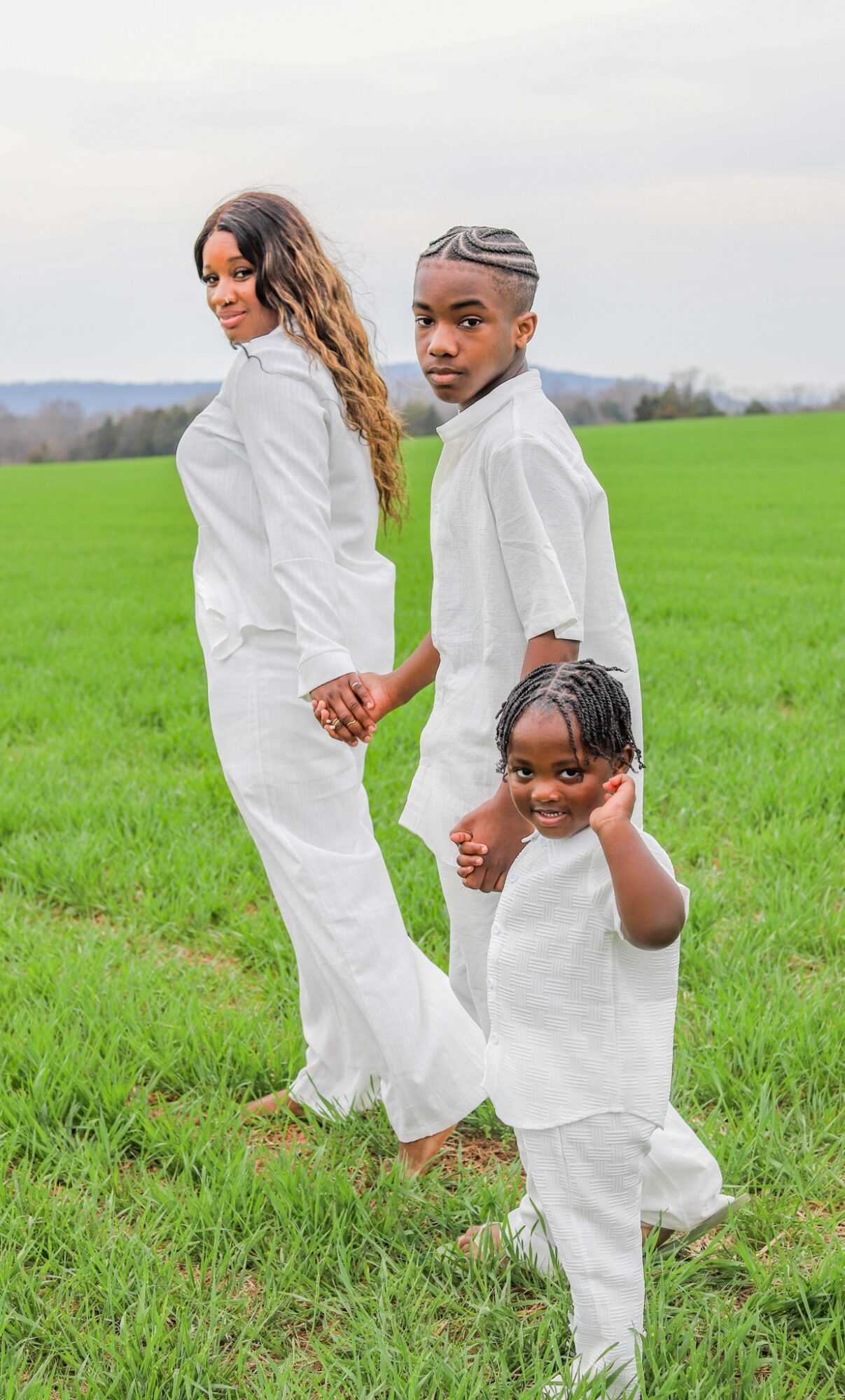 Three people holding hands and smiling in a green field, with a cloudy sky in the background.