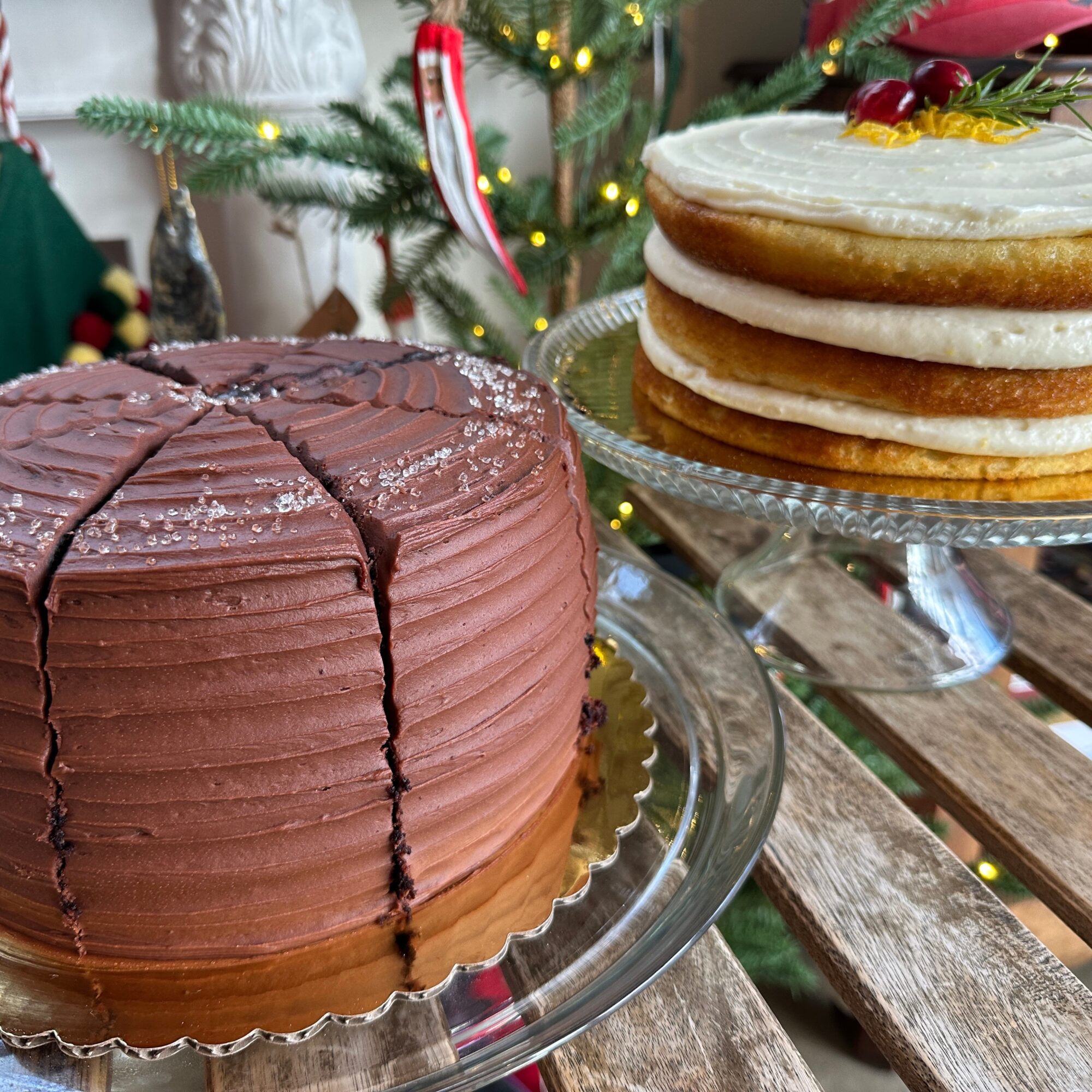Chocolate cake with sliced layers and frosting, and a plate of stacked cookies with icing, on a wooden table with Christmas decorations.