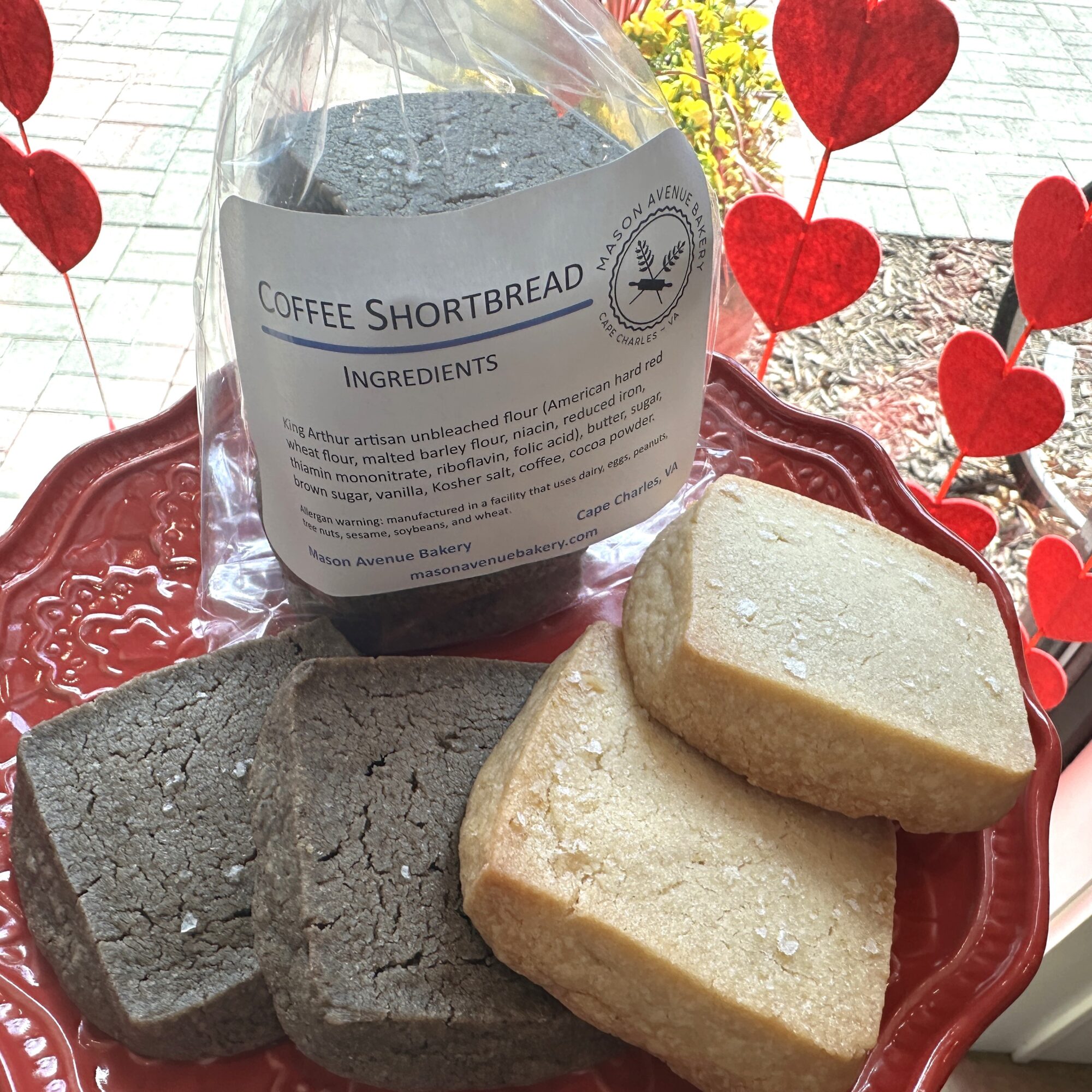 Two shortbread cookies, one light-colored and one darker, on a red decorative tray with a bag labeled 'Coffee Shortbread' behind them, and red heart decorations in the background.