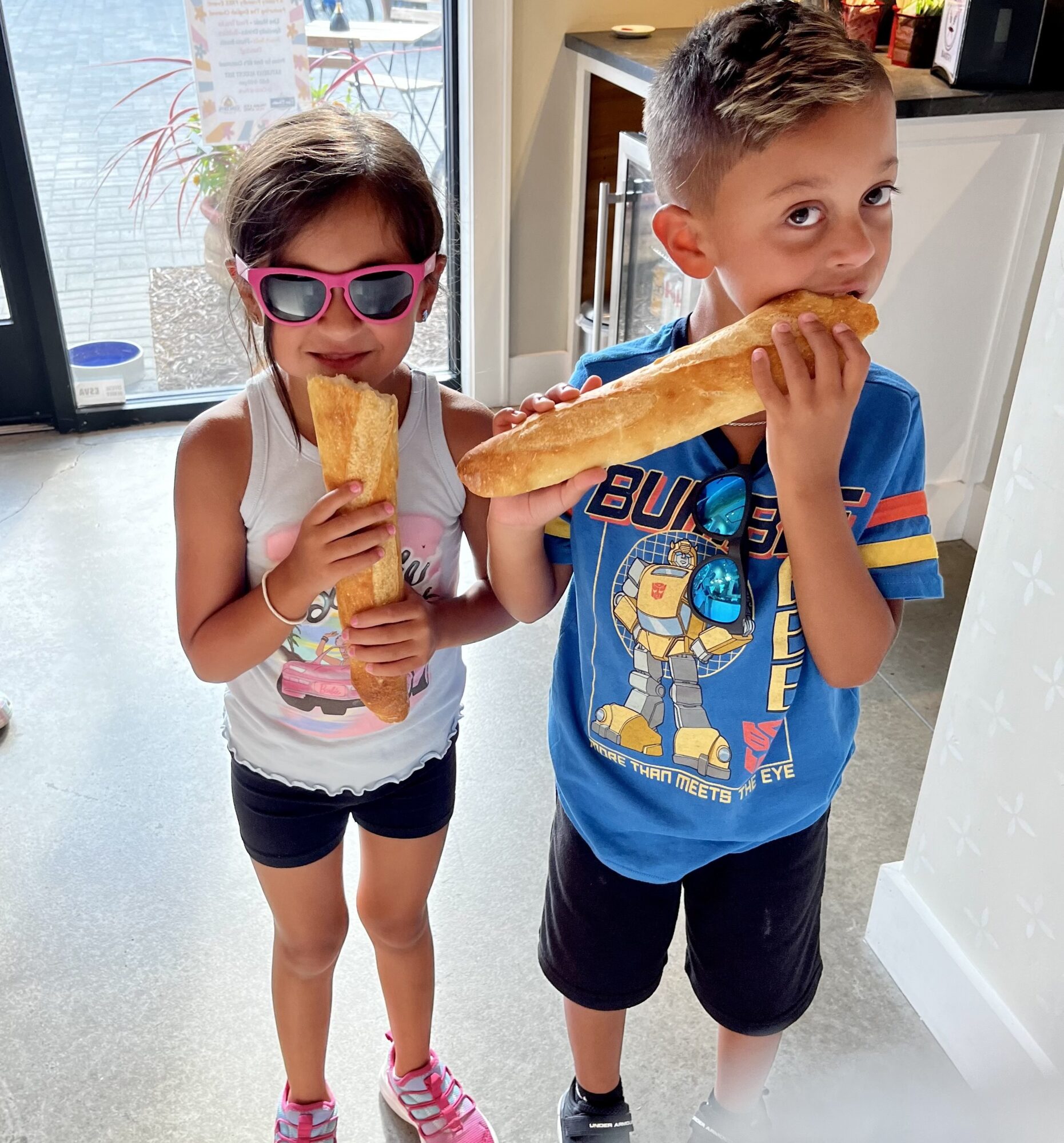 Two children eating baguettes indoors, girl with sunglasses and boy in a blue shirt, near a white cabinet and window.