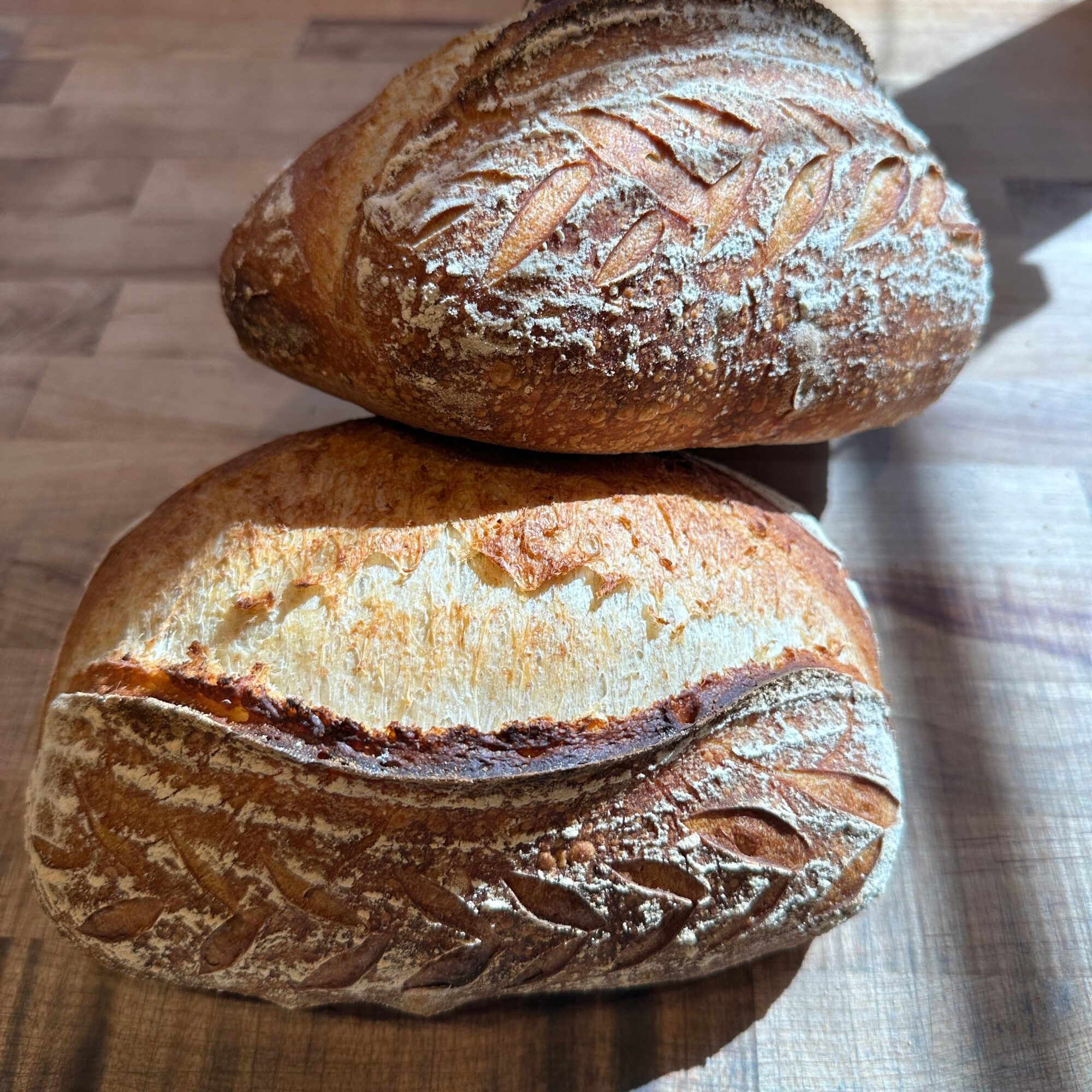 Two loaves of bread on a wooden surface, one with a dusting of flour and decorative scoring on top.