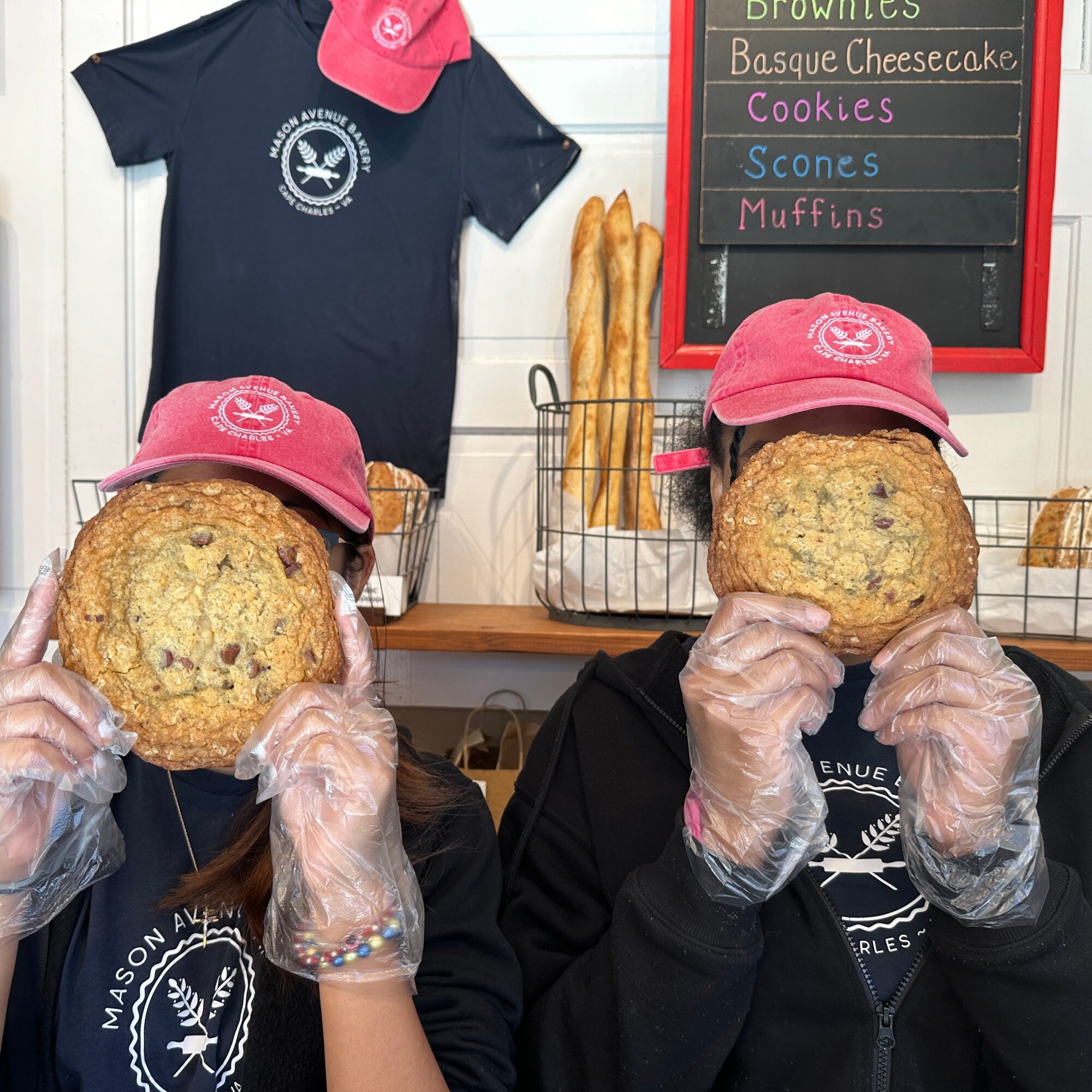Two people wearing pink hats and gloves hold large cookies in front of their faces inside a bakery. A chalkboard menu lists baked goods.
