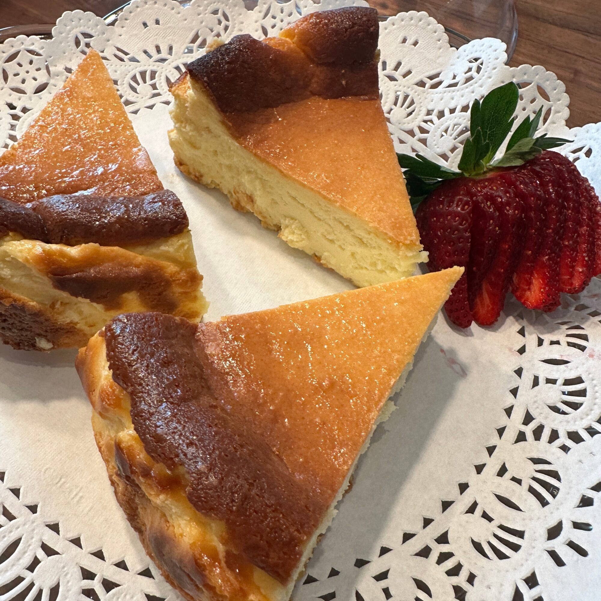 Three slices of cheesecake with a strawberry on a decorative paper doily, on a wooden surface.