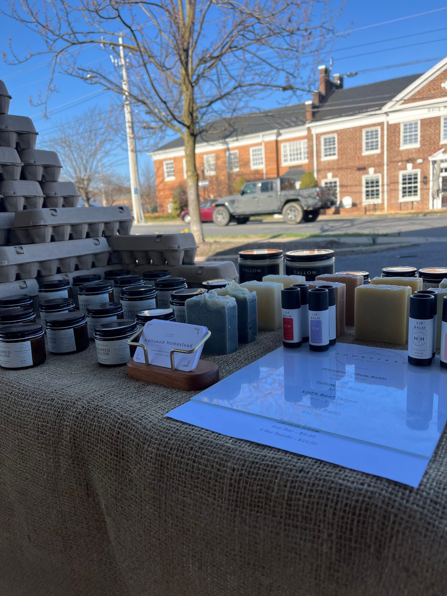 Table with various bottles, jars, and a sheet of paper outdoors, with buildings and a tree in the background.