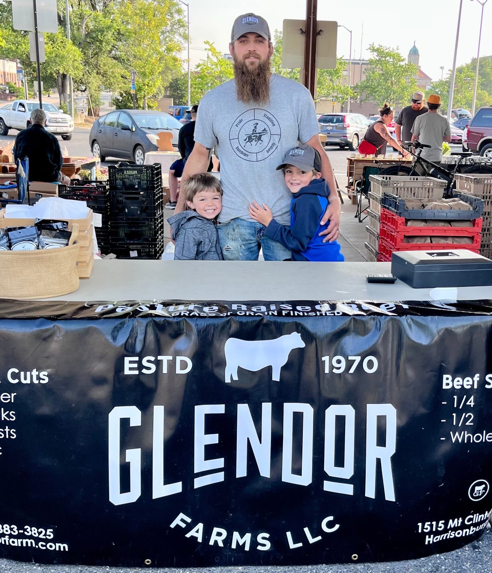 Man with beard and cap standing behind two children at outdoor market stall, with cars and people in background.