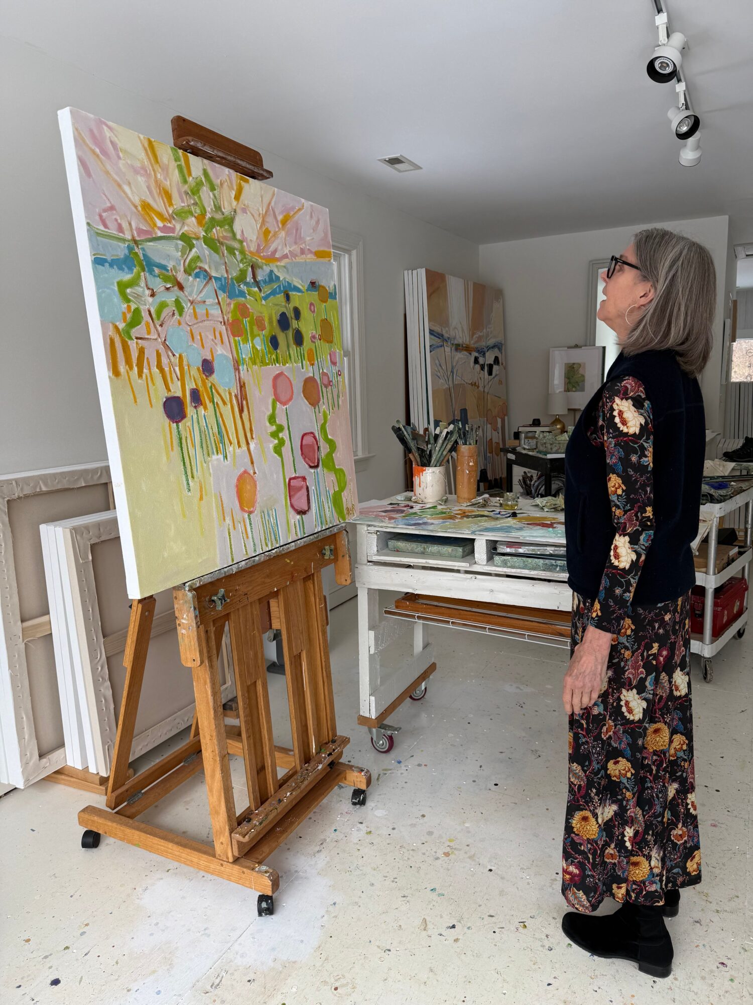 Woman with gray hair wearing a floral dress and black cardigan looking at a colorful painting on an easel in an art studio.