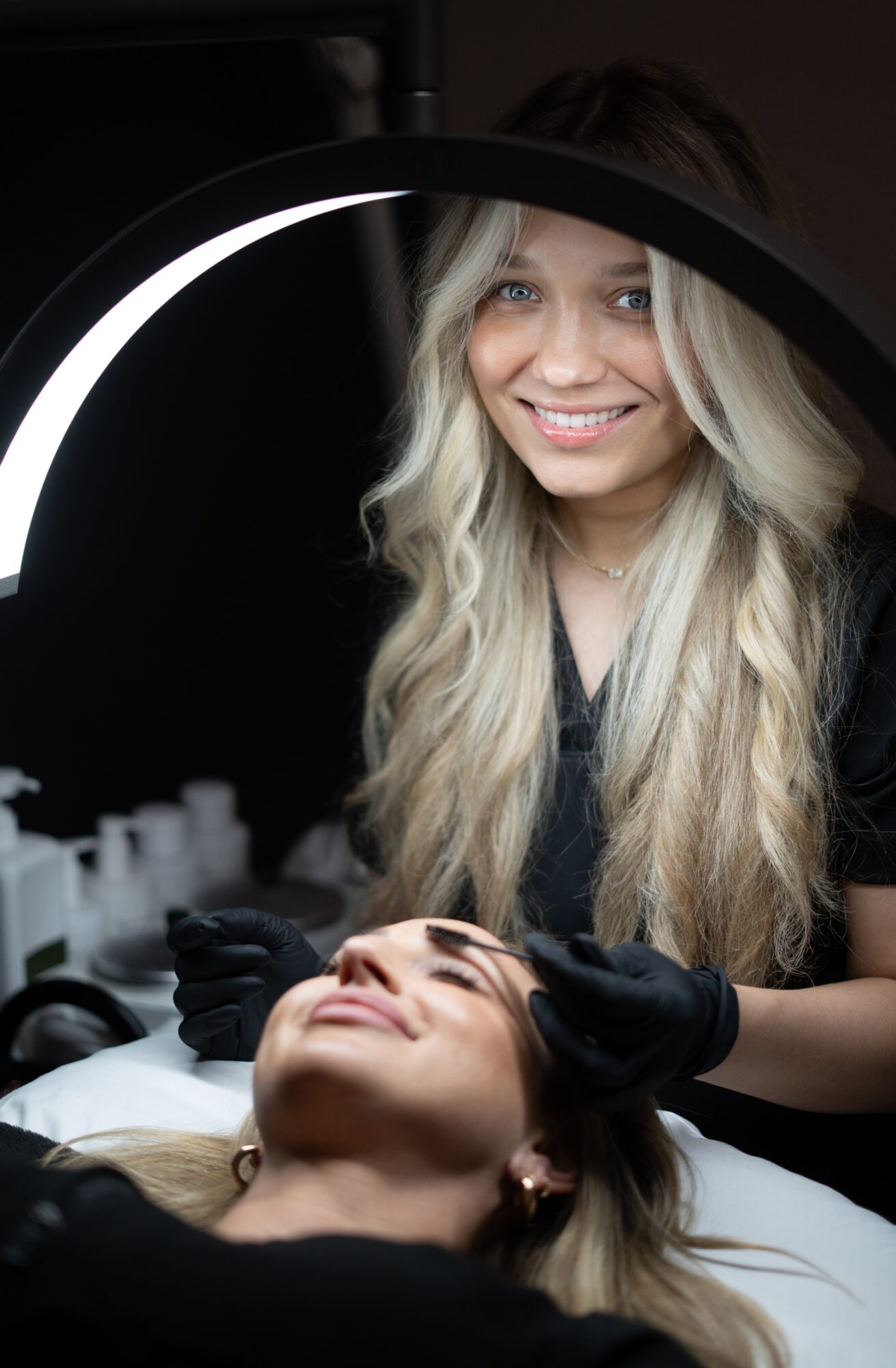 A woman with long blonde hair smiling while receiving a facial treatment, with a beauty ring light in the foreground.