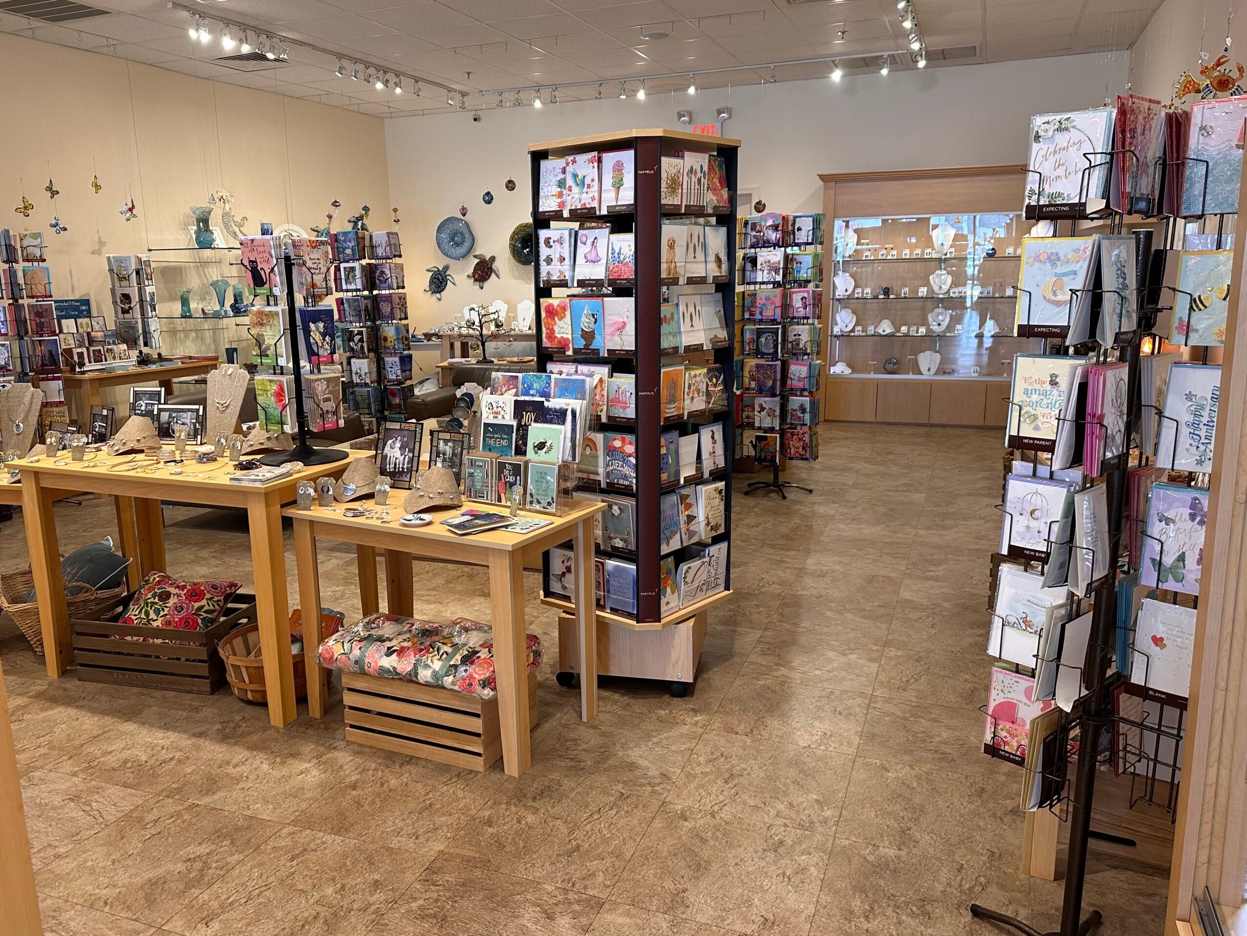 Interior of a store with greeting cards and gift items on display, wooden tables, and shelves, with a window in the background.
