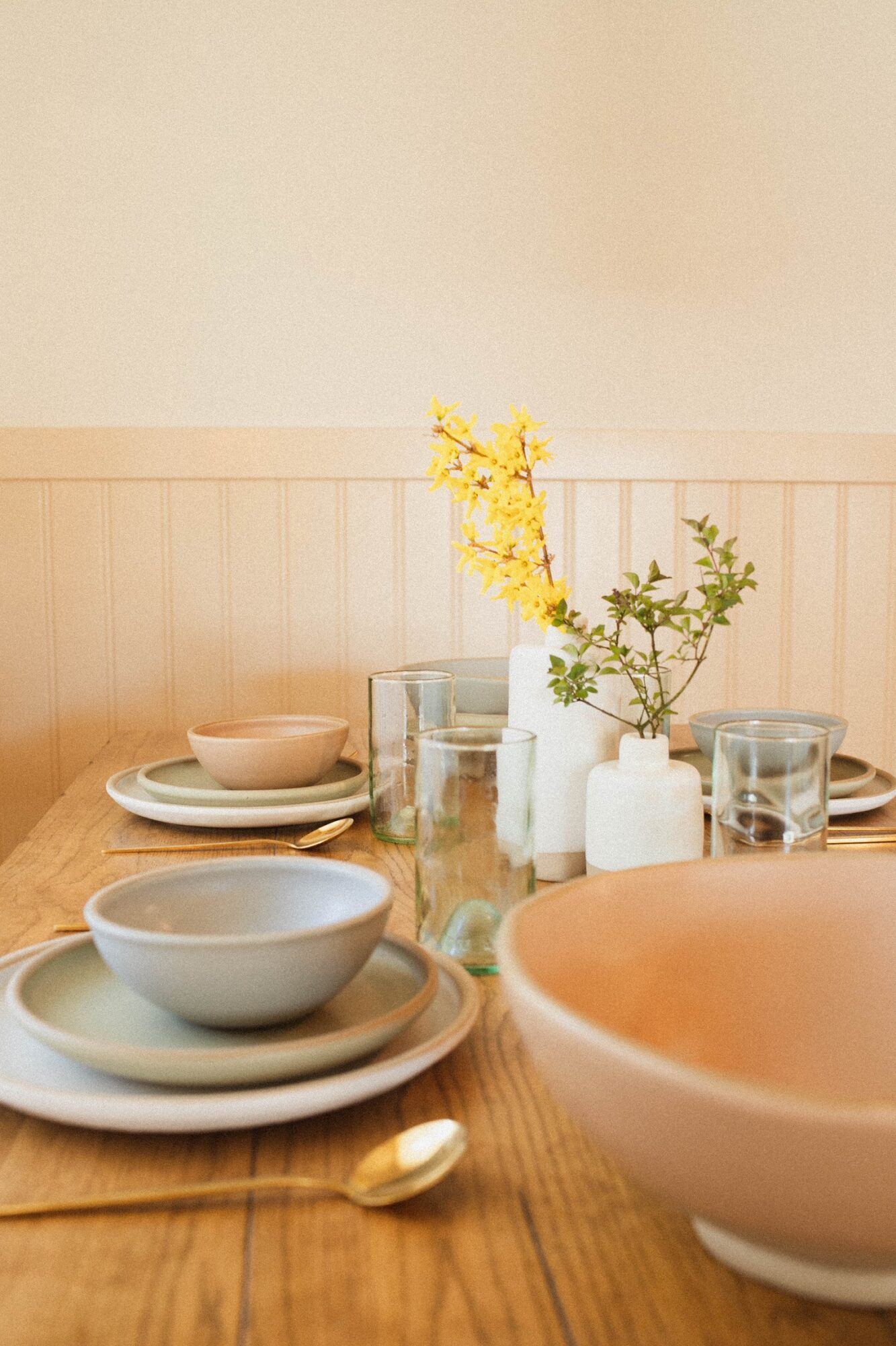 Table setting with bowls, glasses, and a vase with yellow flowers on a wooden table, against a light wall.