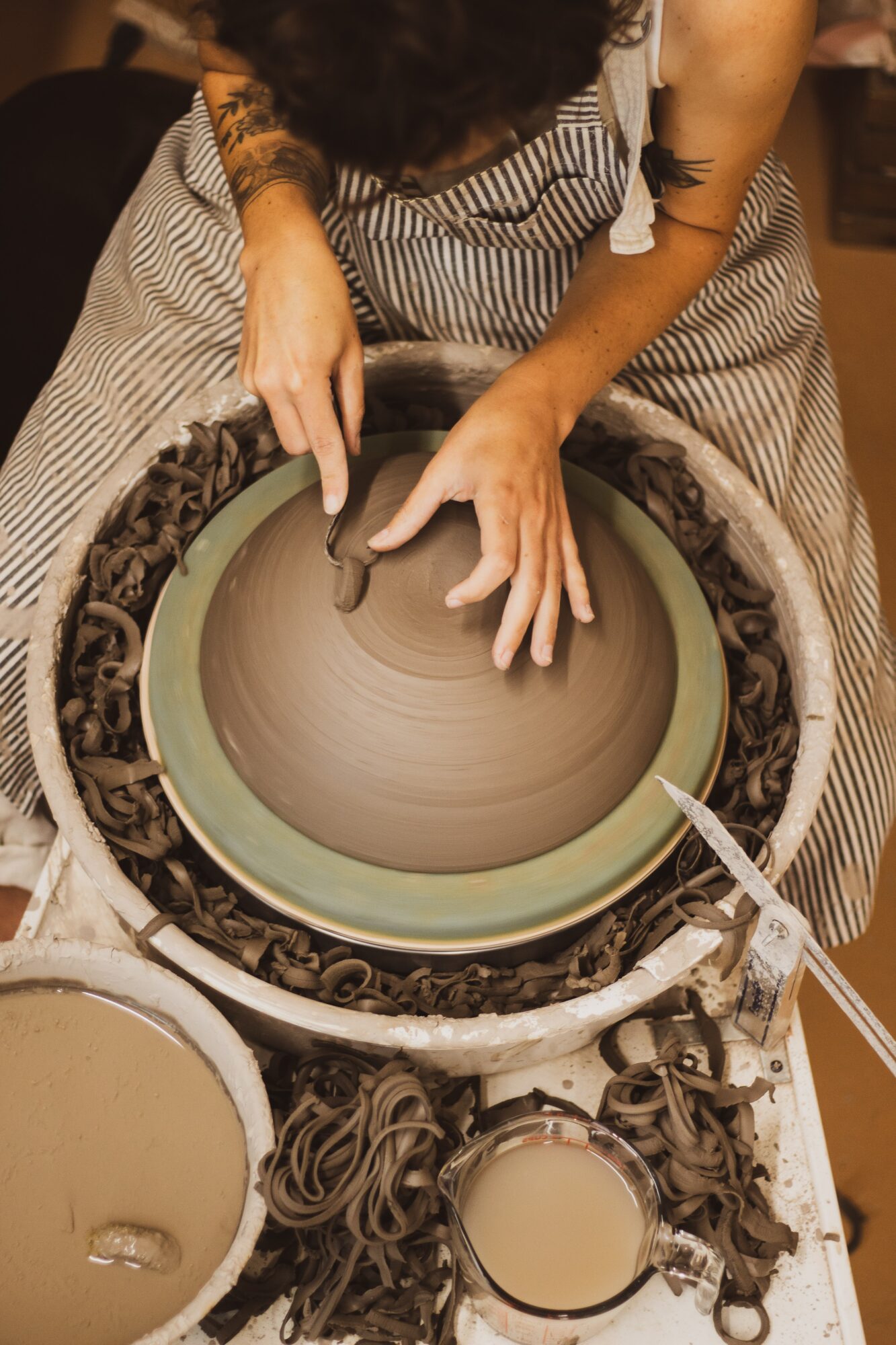 Person shaping clay on a pottery wheel with tools, surrounded by clay and pottery supplies, viewed from above.
