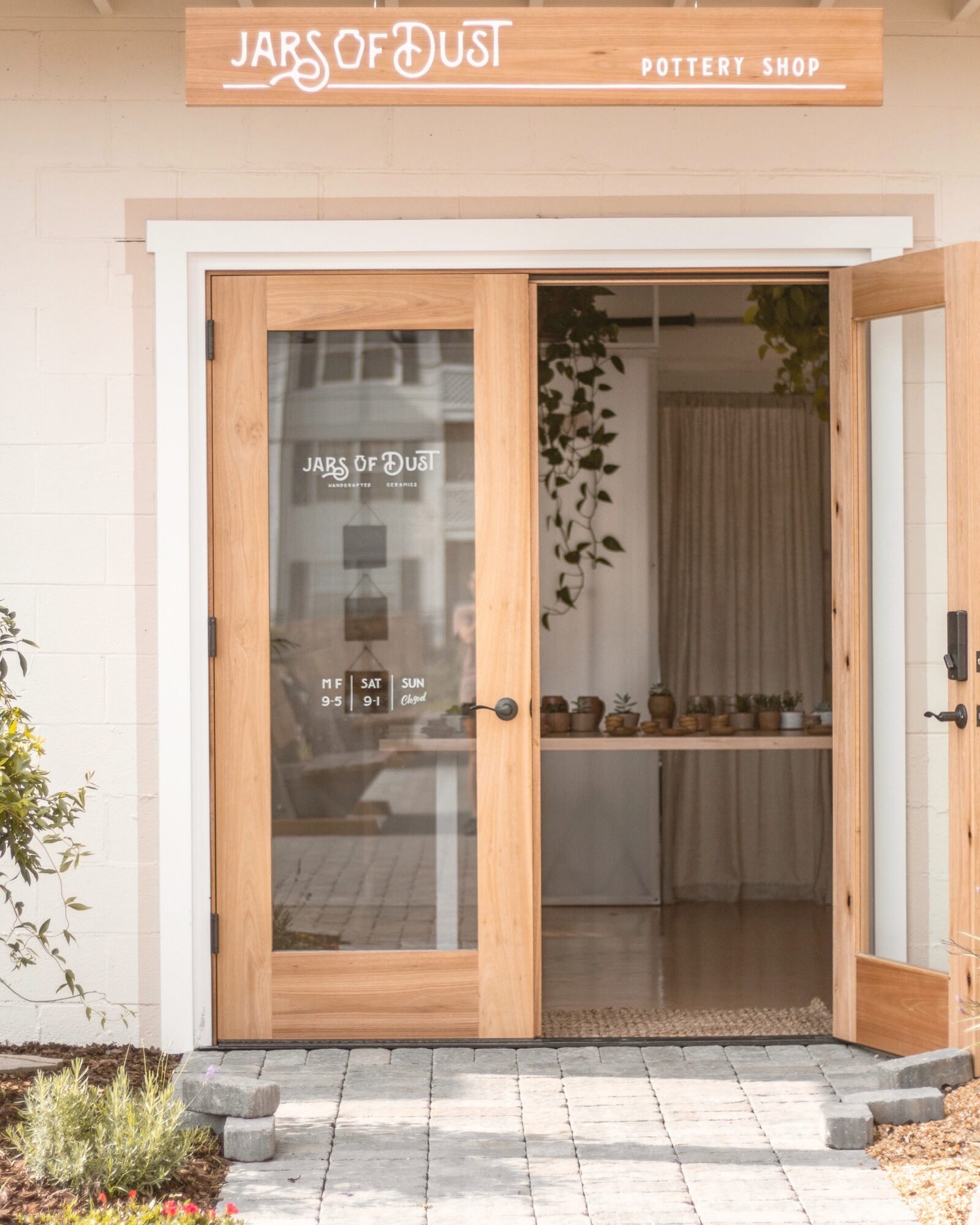 Wooden sliding door with glass panels leading into a shop, sign above reads 'Jars of Dust Pottery Shop', plants outside.
