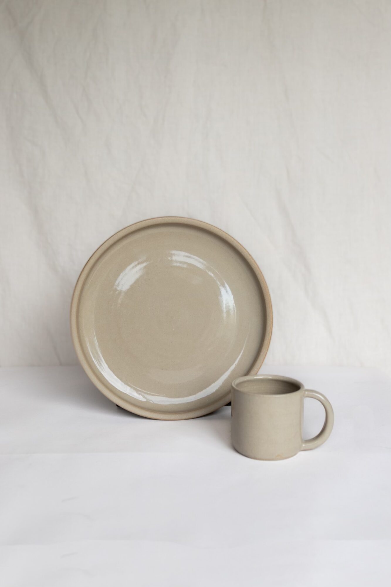 A beige plate and a matching mug are placed on a white surface against a white fabric background.