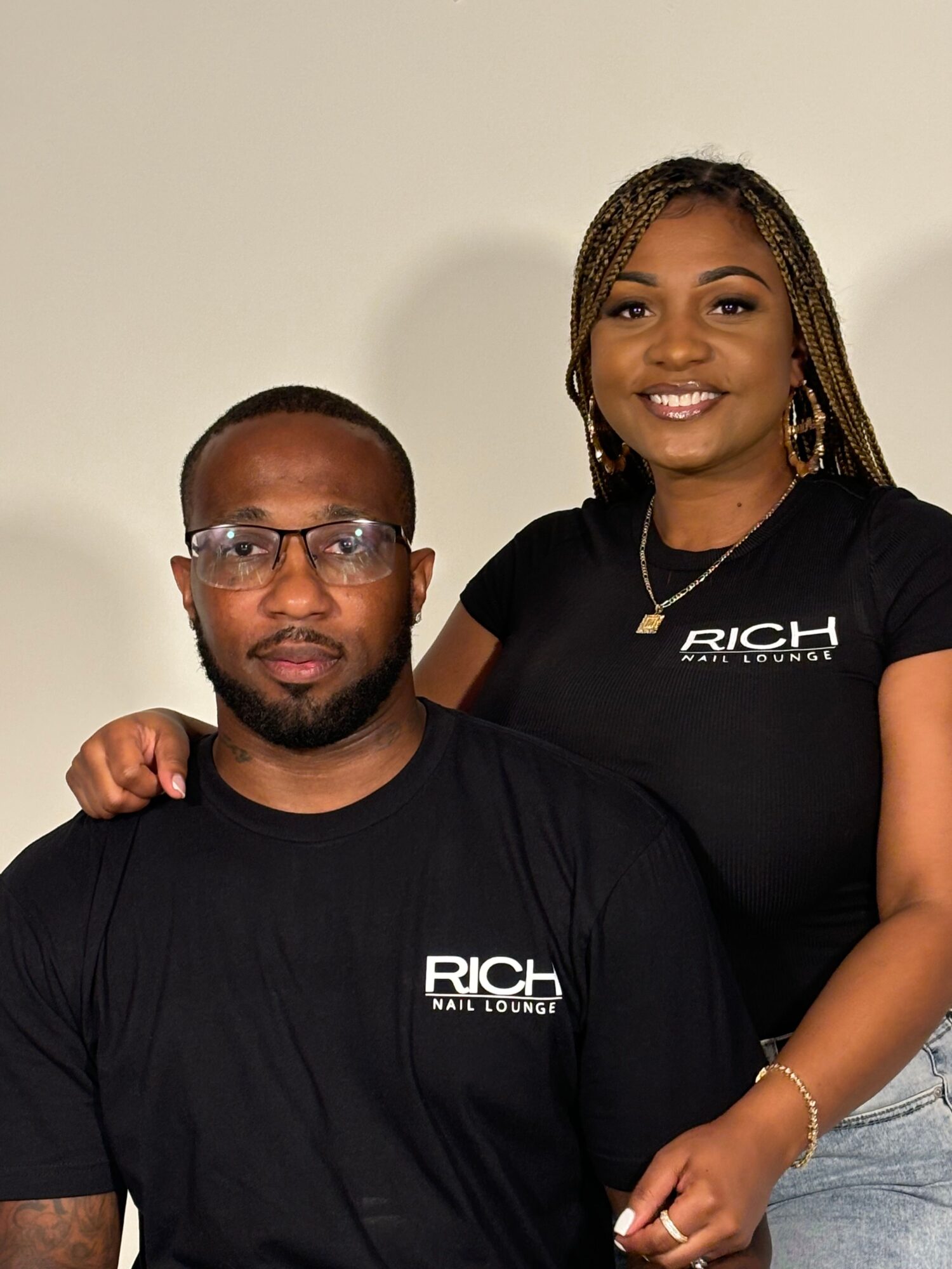 A woman and a man pose together, both wearing black shirts with the word RICH printed on them, smiling at the camera.