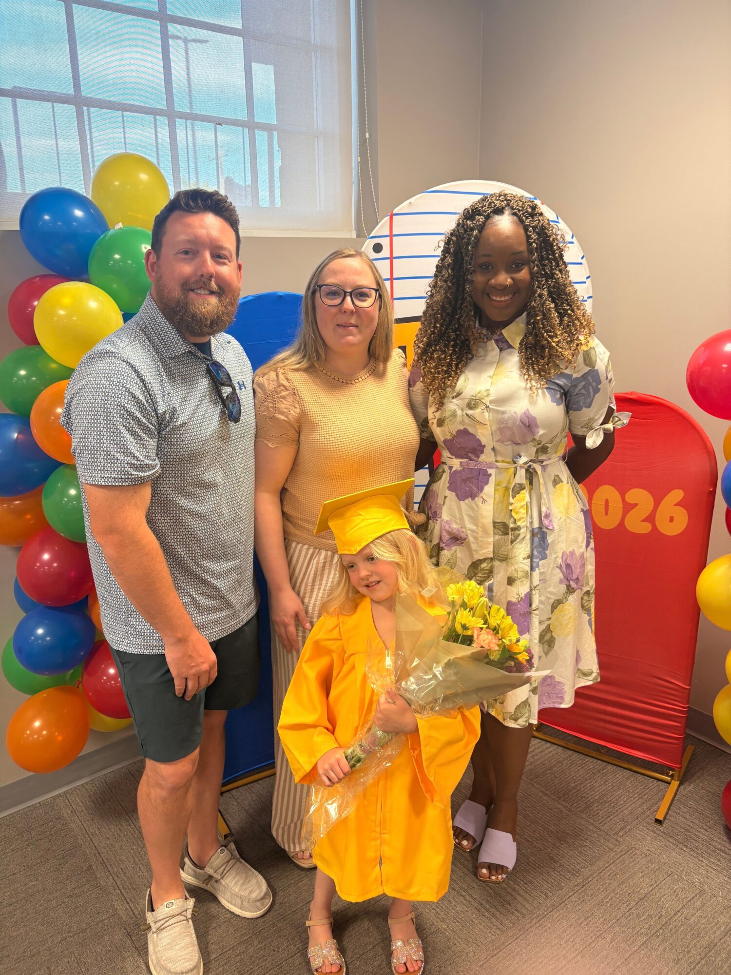 Three people standing indoors with colorful balloons and a red sign in the background, smiling and holding flowers.