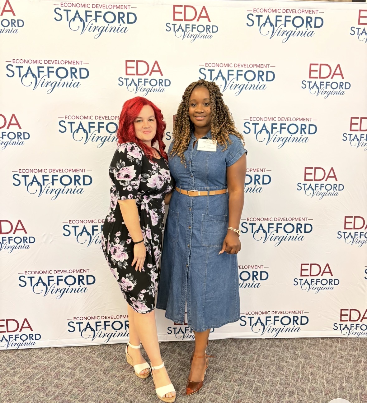 Two women standing in front of a backdrop with logos, smiling, one with red hair and floral dress, the other with curly hair and denim dress.
