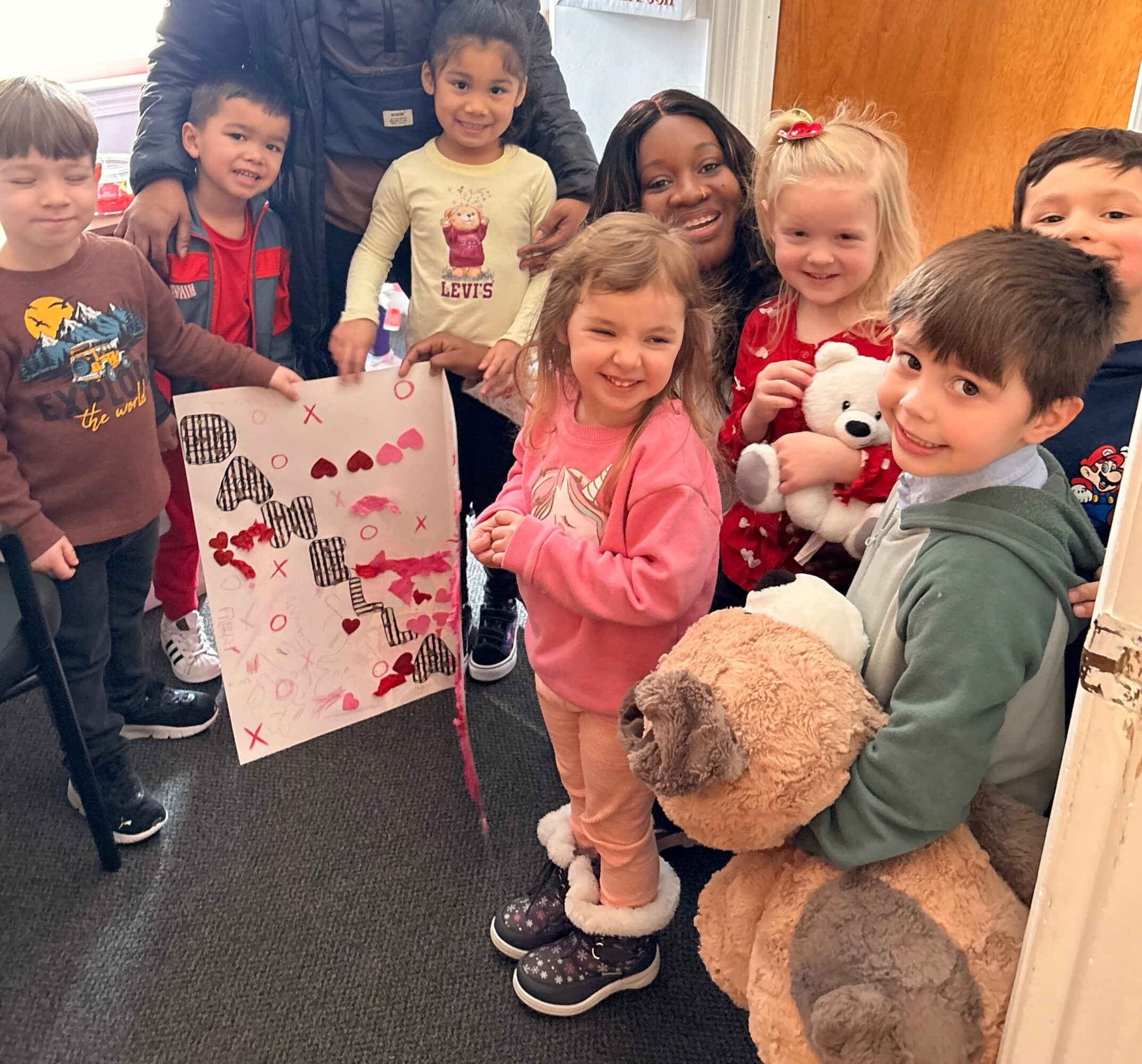 Group of children and an adult smiling, holding gifts and a large teddy bear, in a room with wooden door.
