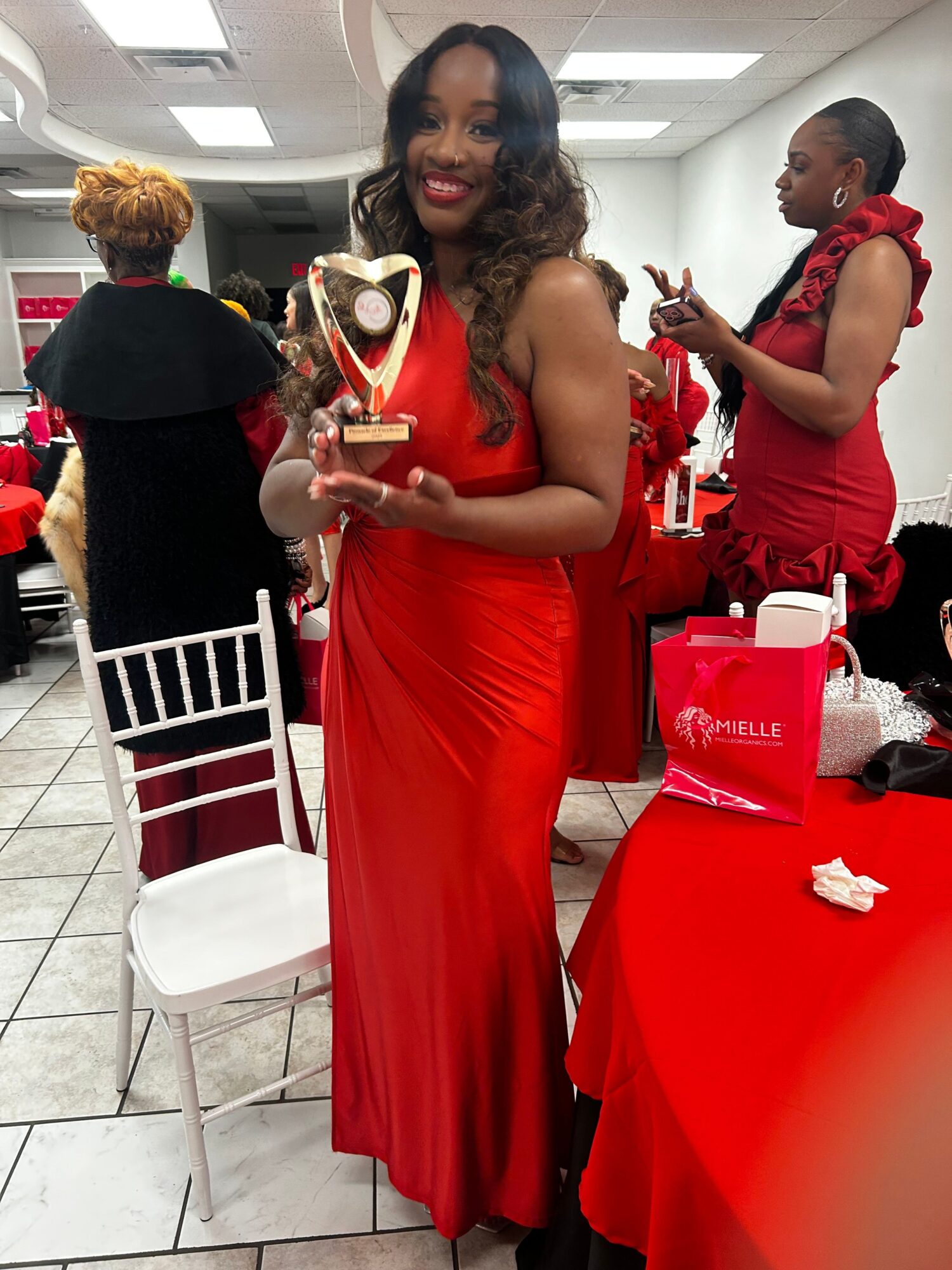 Two women in red dresses at a table with drinks and decorations, one holding a gift, in a festive setting.