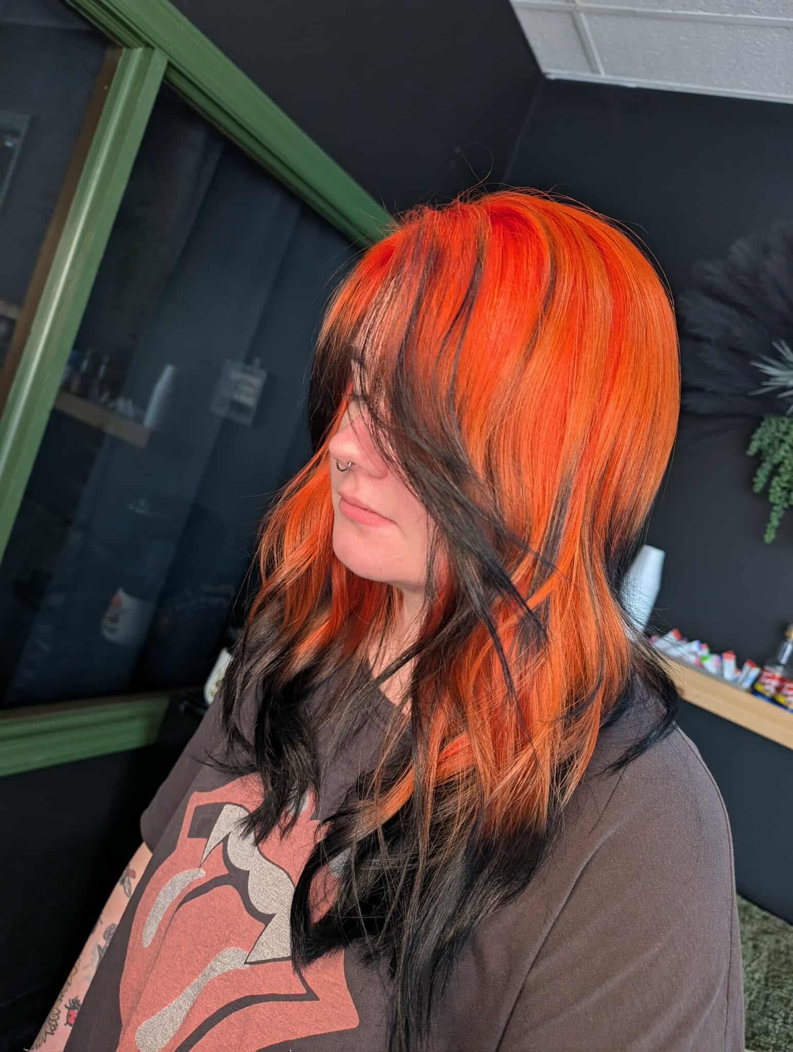 Woman with orange and black hair looking down, indoors with green window frame and dark wall behind her.