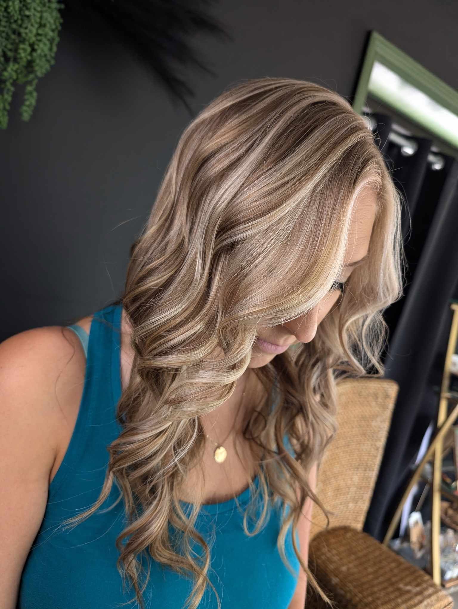 Side view of a woman with long, wavy blonde hair wearing a blue top, indoors with dark walls and furniture.