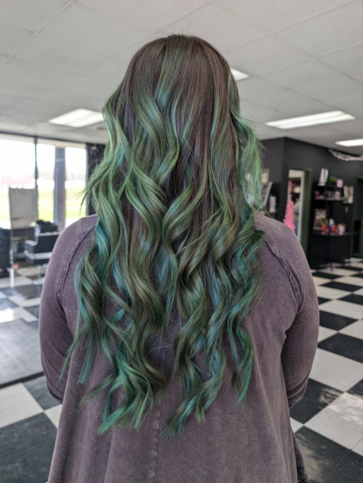 Back view of a person with long, wavy hair dyed in shades of green and blue, indoors with checkered floor.