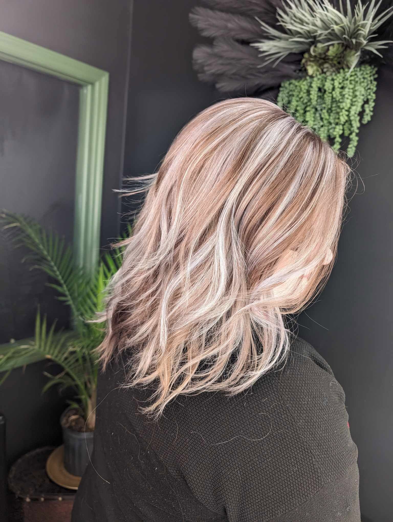 Person with shoulder-length wavy blonde hair, facing away, in a room with plants and dark wall.