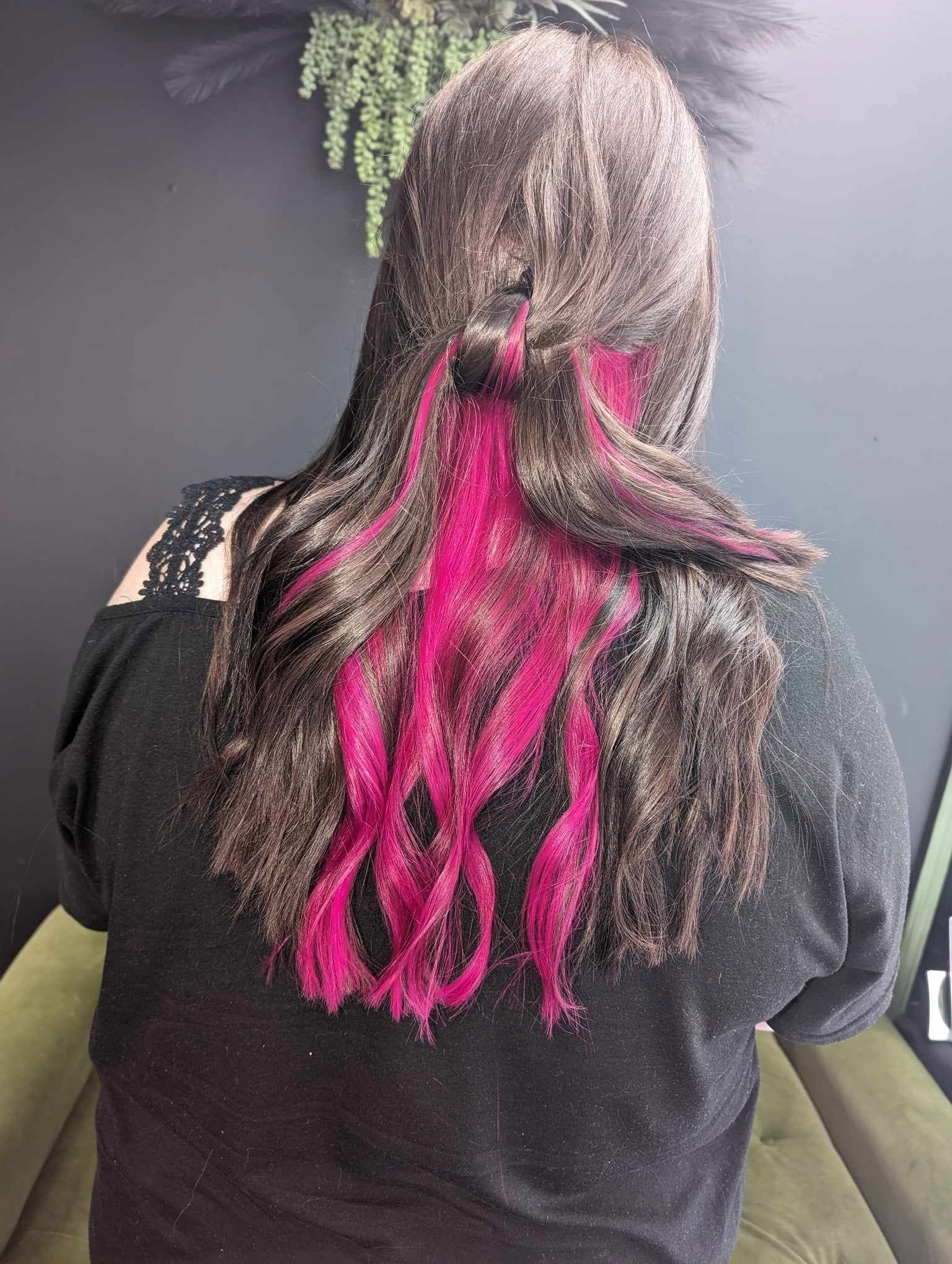 Back of a person with long wavy hair, half dyed bright pink, wearing a black top, against a dark background.