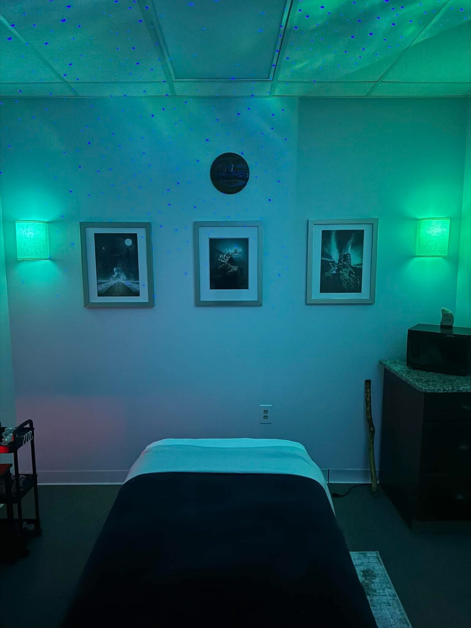 Room with three framed pictures on the wall, massage table in the center, and green lighting, creating a calming atmosphere.