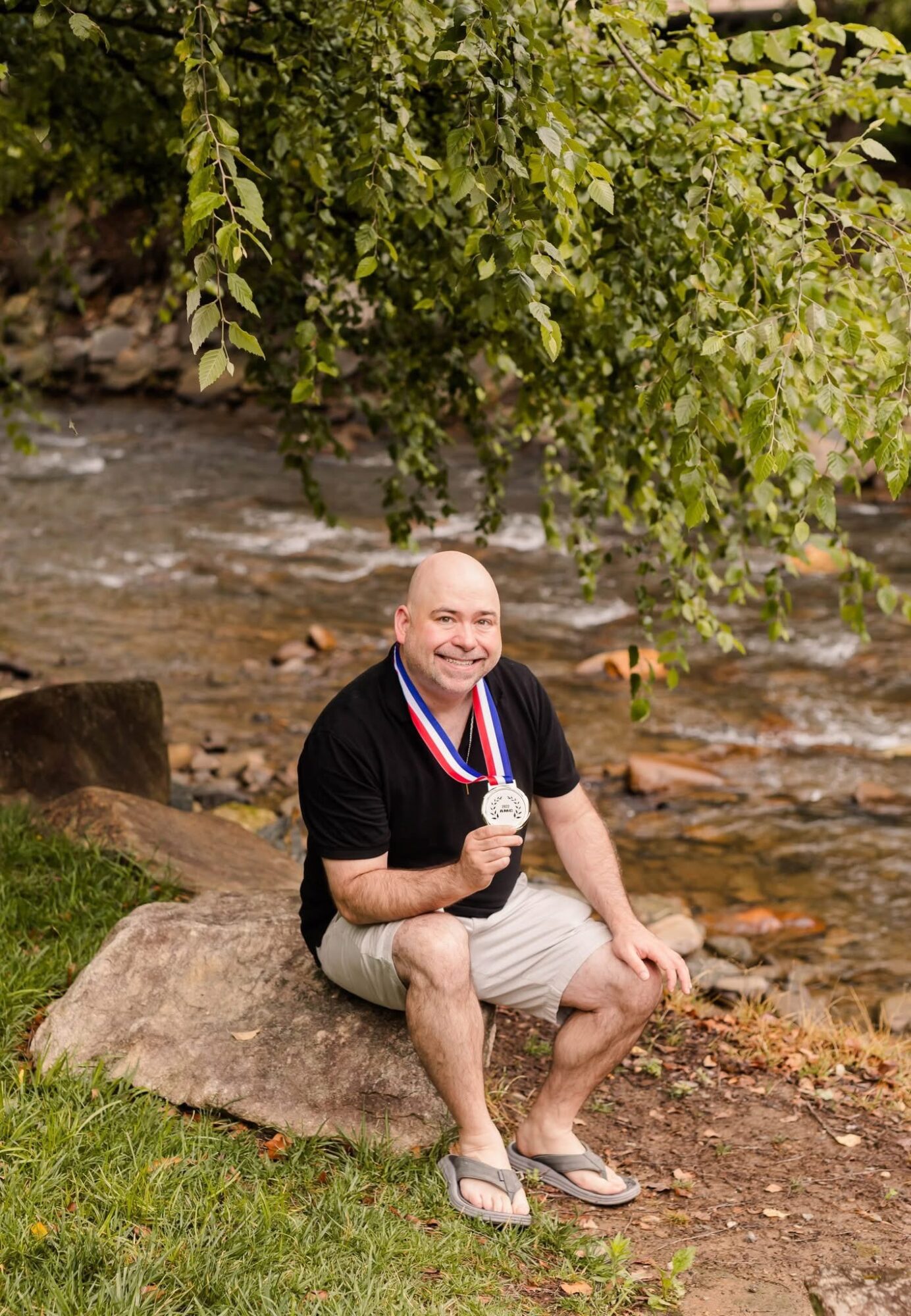 Man sitting on a rock near a river, holding a medal, smiling, with green trees overhead.