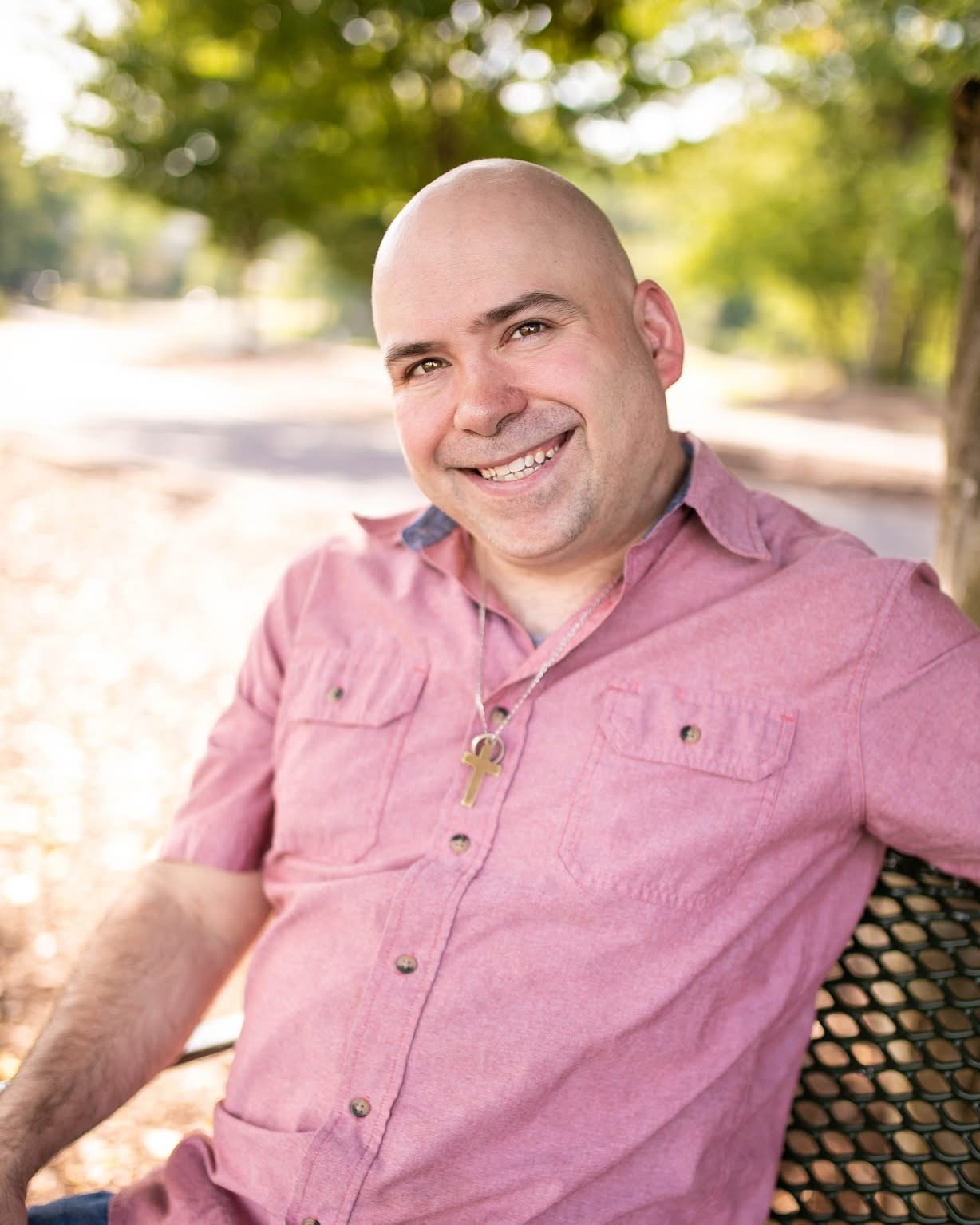 Smiling man with a shaved head wearing a pink shirt sitting outdoors on a bench, trees in background.