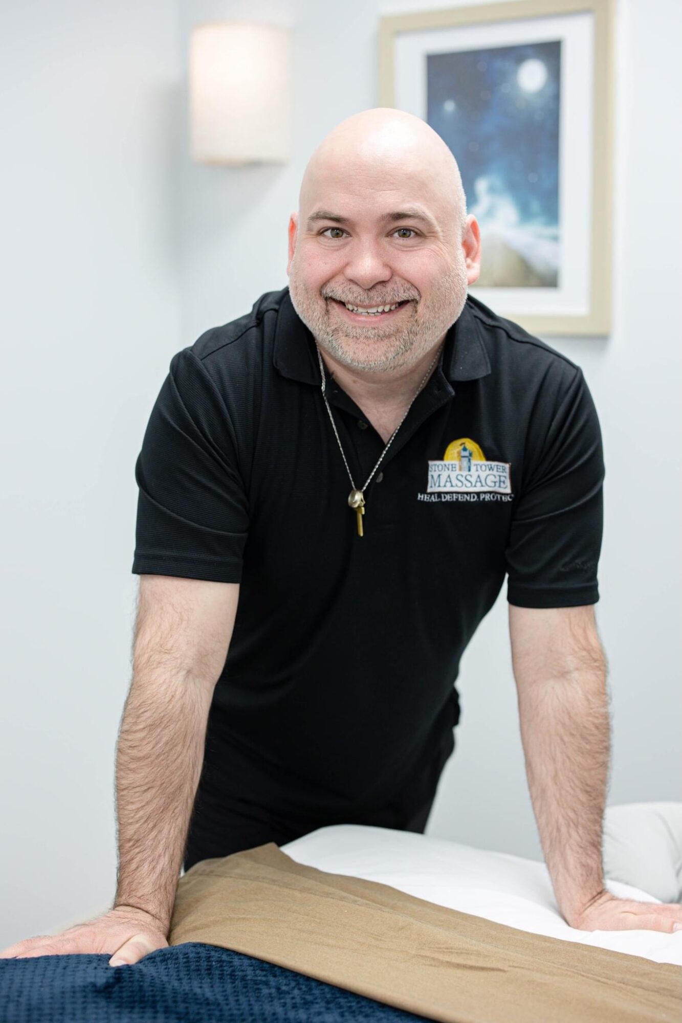 Smiling man with a shaved head leaning on a massage table in a room with a framed picture and a lamp.
