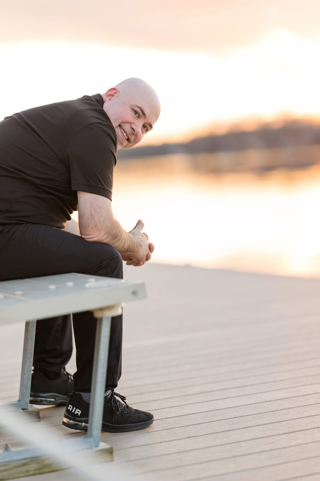 Person with short white hair sitting on a bench outdoors near water, smiling at the camera during sunset.